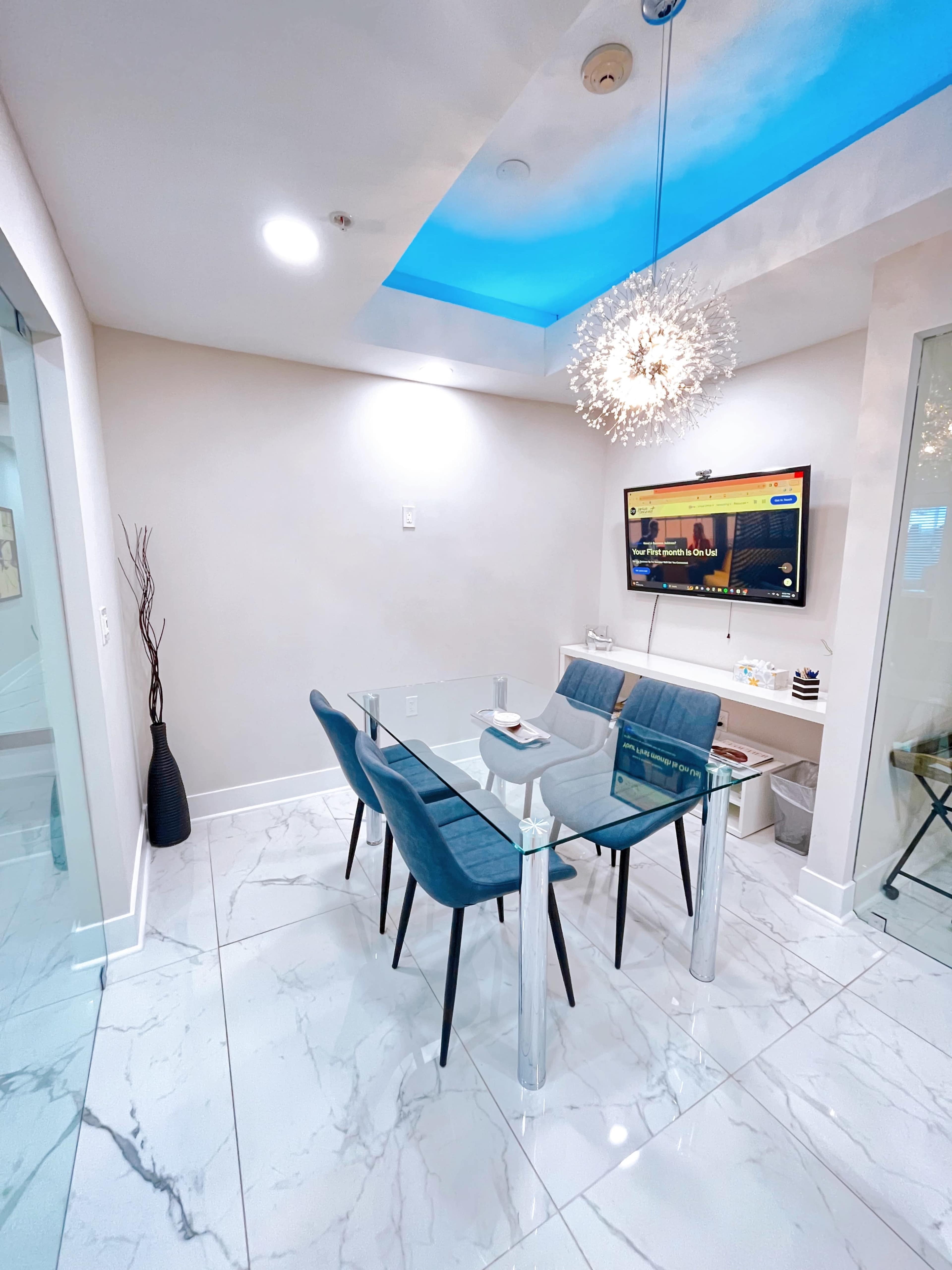 A modern dining area features a glass table surrounded by four blue chairs, with a television mounted on the wall and a decorative light fixture above.