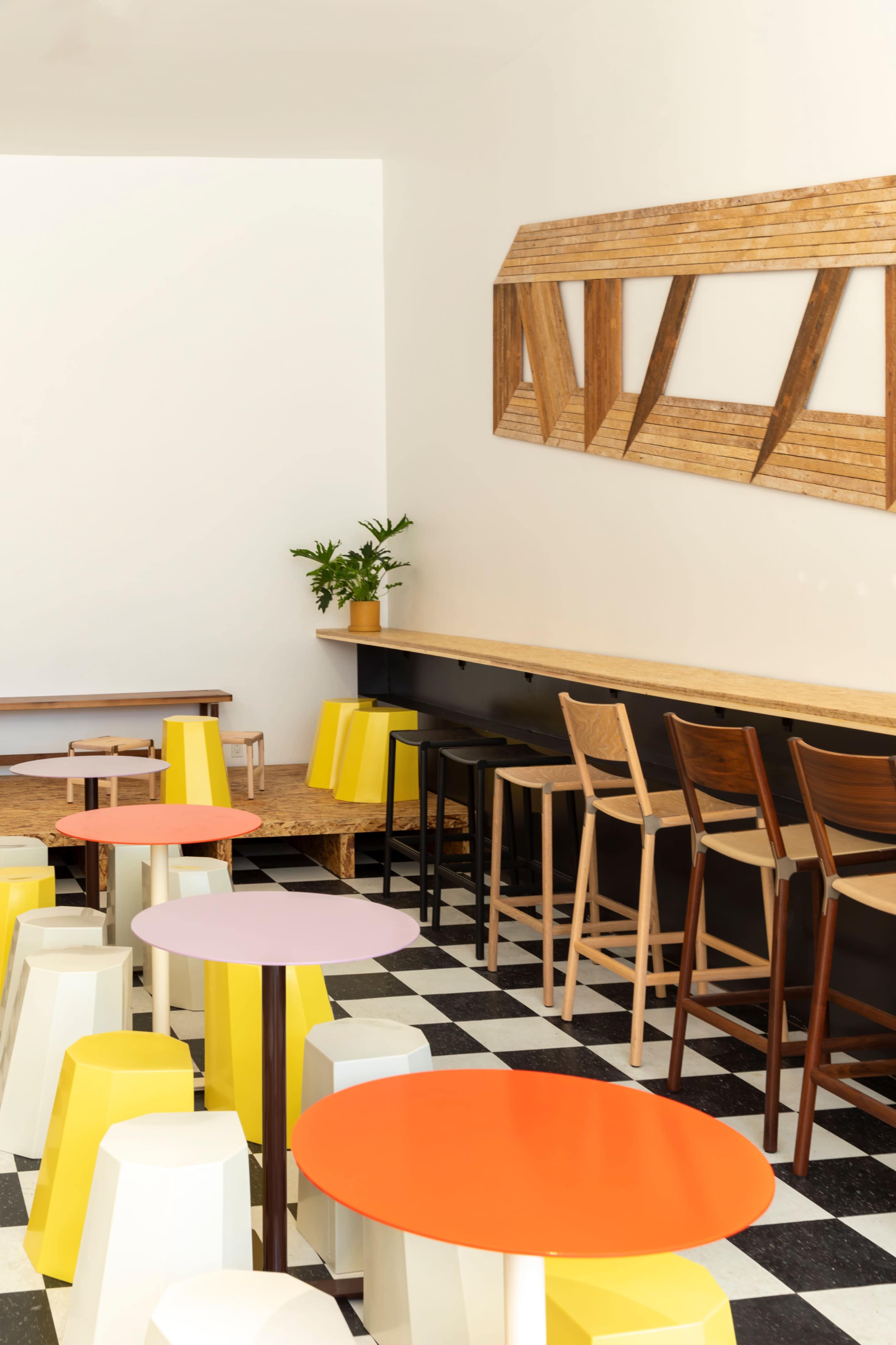 The image shows a modern café interior with colorful stools and tables, featuring a mix of wood and painted surfaces against a black and white checkered floor.