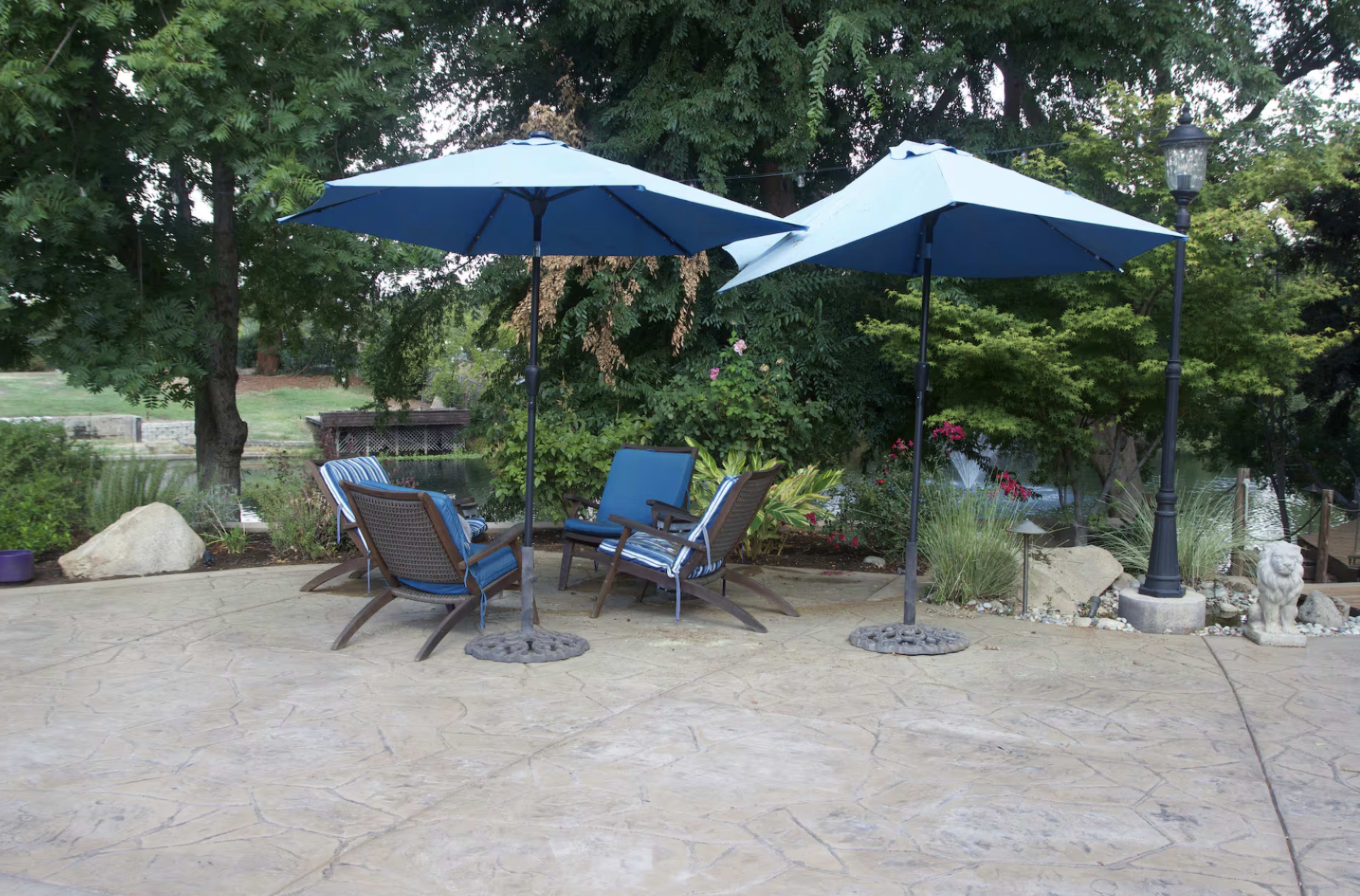 The image shows a patio area with two blue umbrellas over a seating arrangement of four chairs and a table, surrounded by greenery and decorative elements.