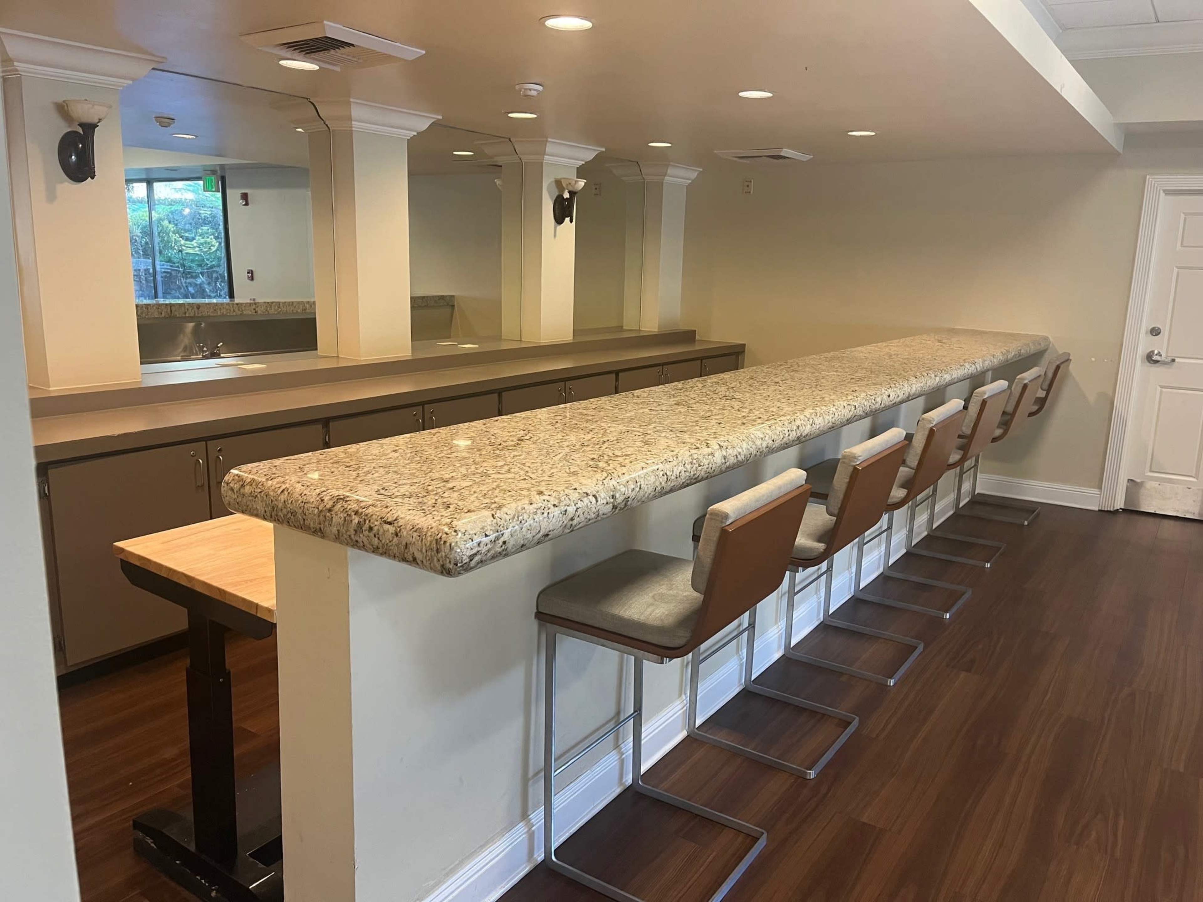 A long, granite countertop with a row of metal and wood bar stools in an indoor space, illuminated by overhead lighting and facing a wall of mirrors.