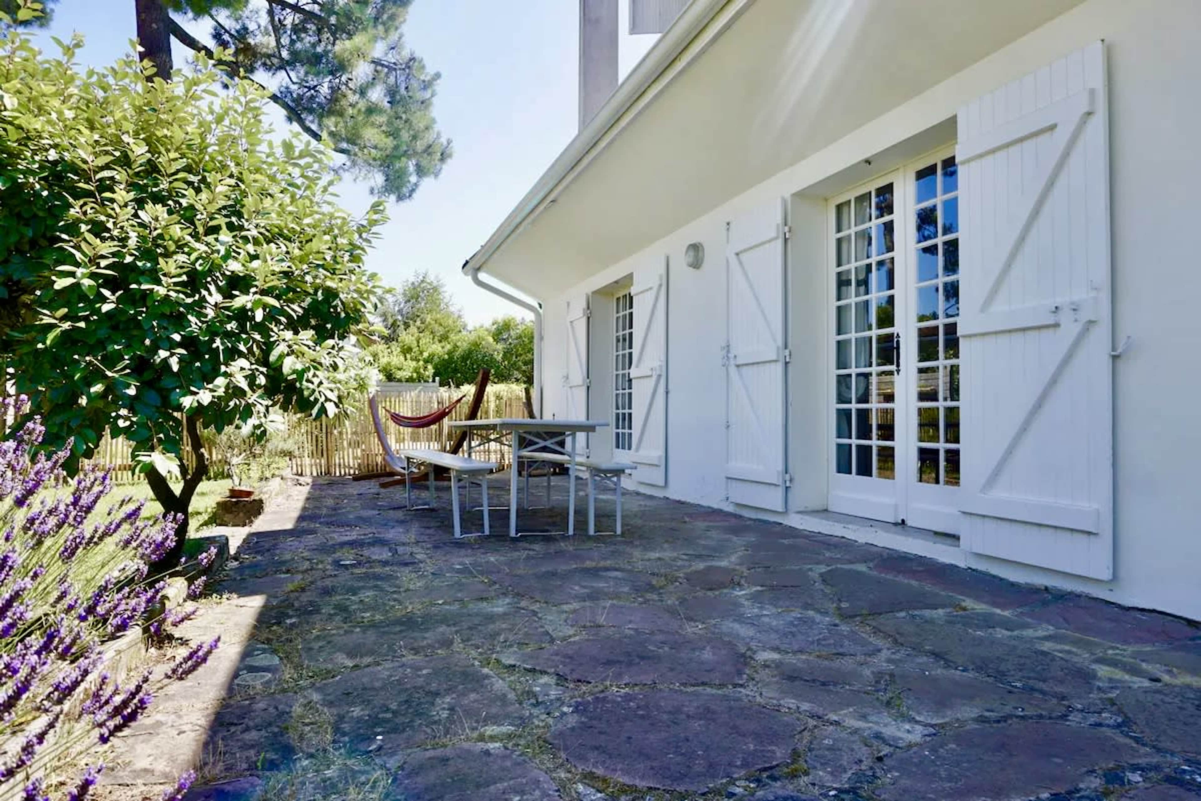 A stone-paved patio is adorned with a table and chairs, surrounded by greenery and a hammock attached to a tree.