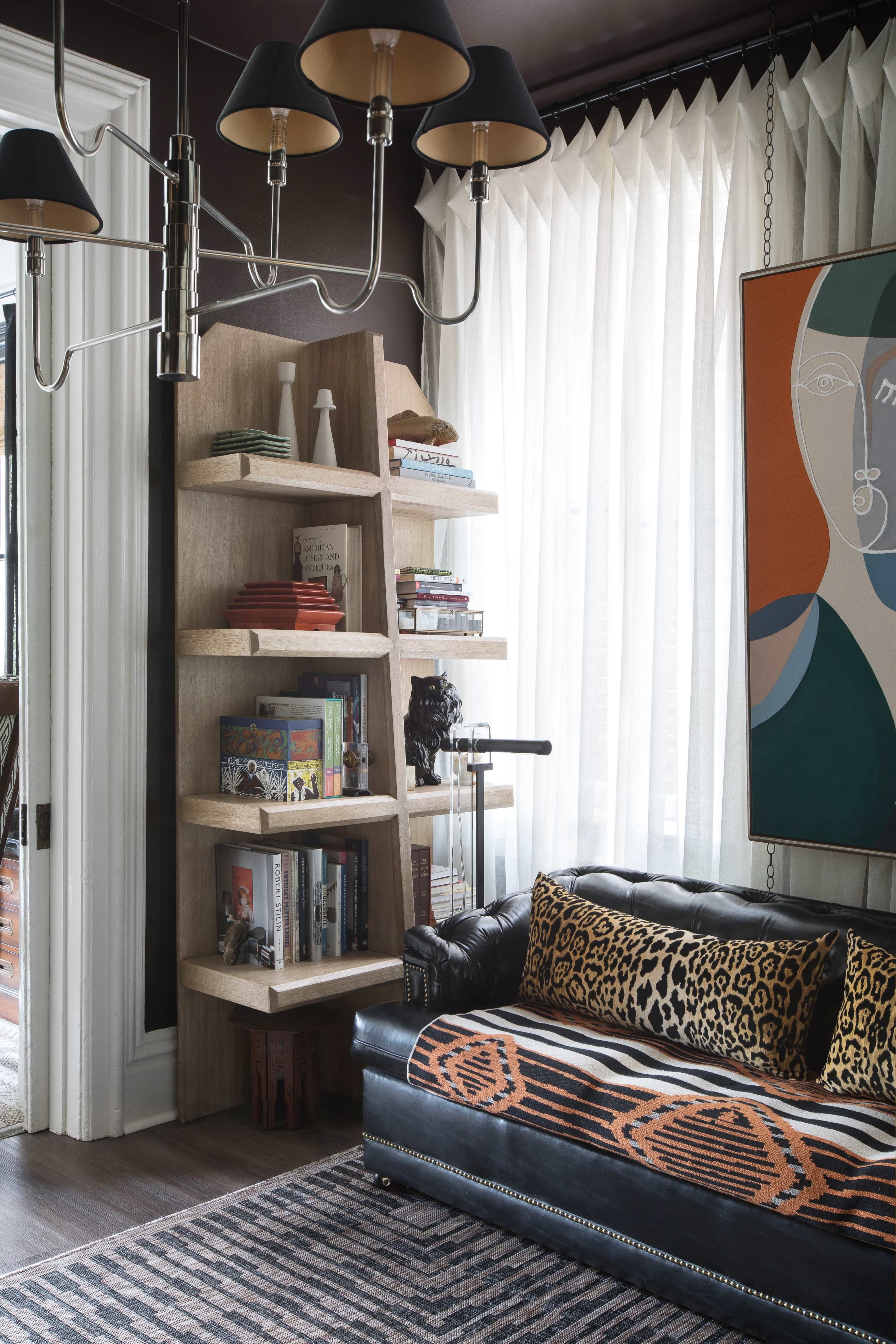 A black leather sofa with a patterned throw pillow is positioned next to a wooden bookshelf filled with books and decor, beneath a chandelier and large windows with sheer curtains.