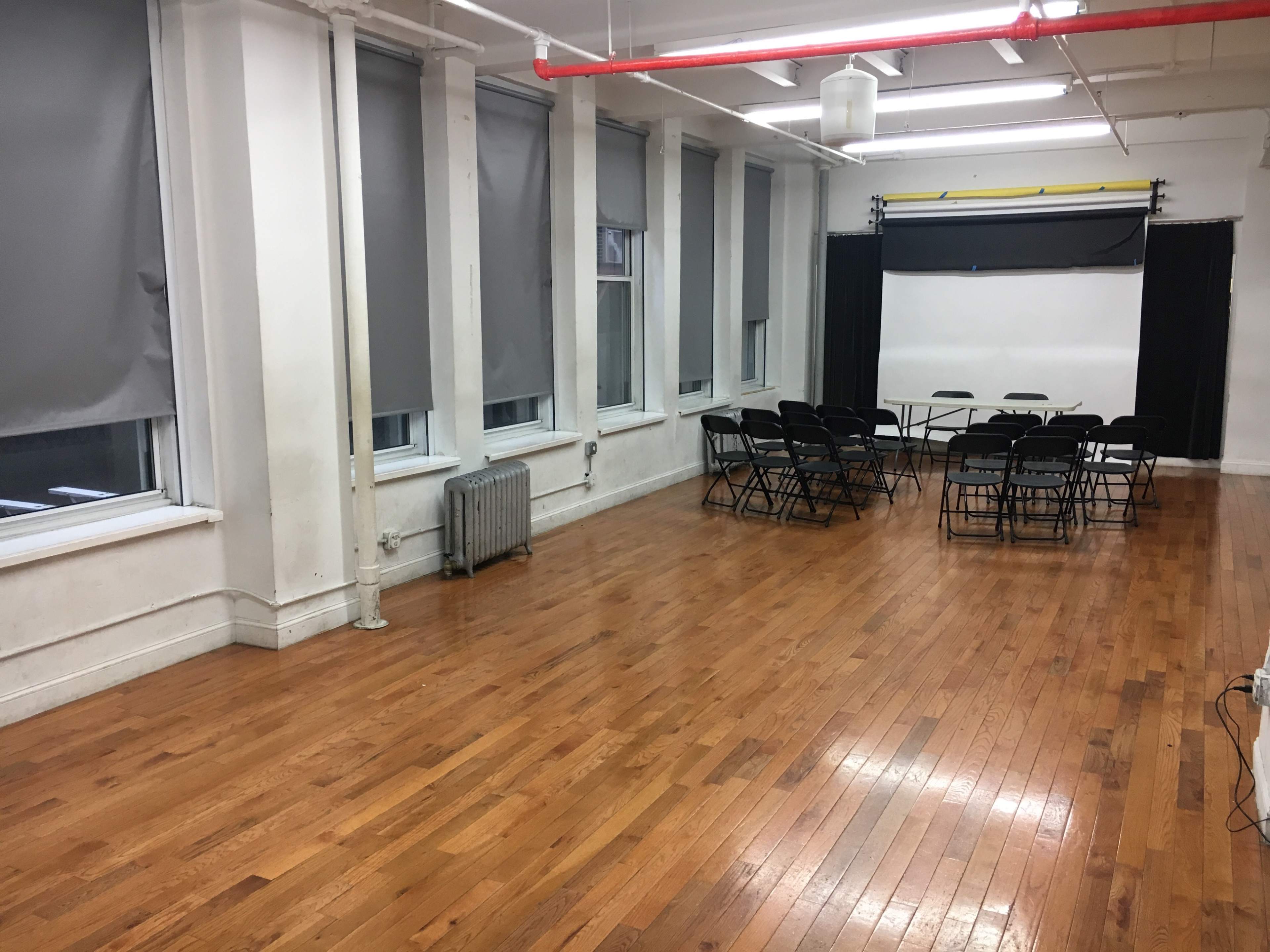 The image shows a large, empty room with hardwood floors, gray blinds, and a setup of black chairs arranged around a table at the back.