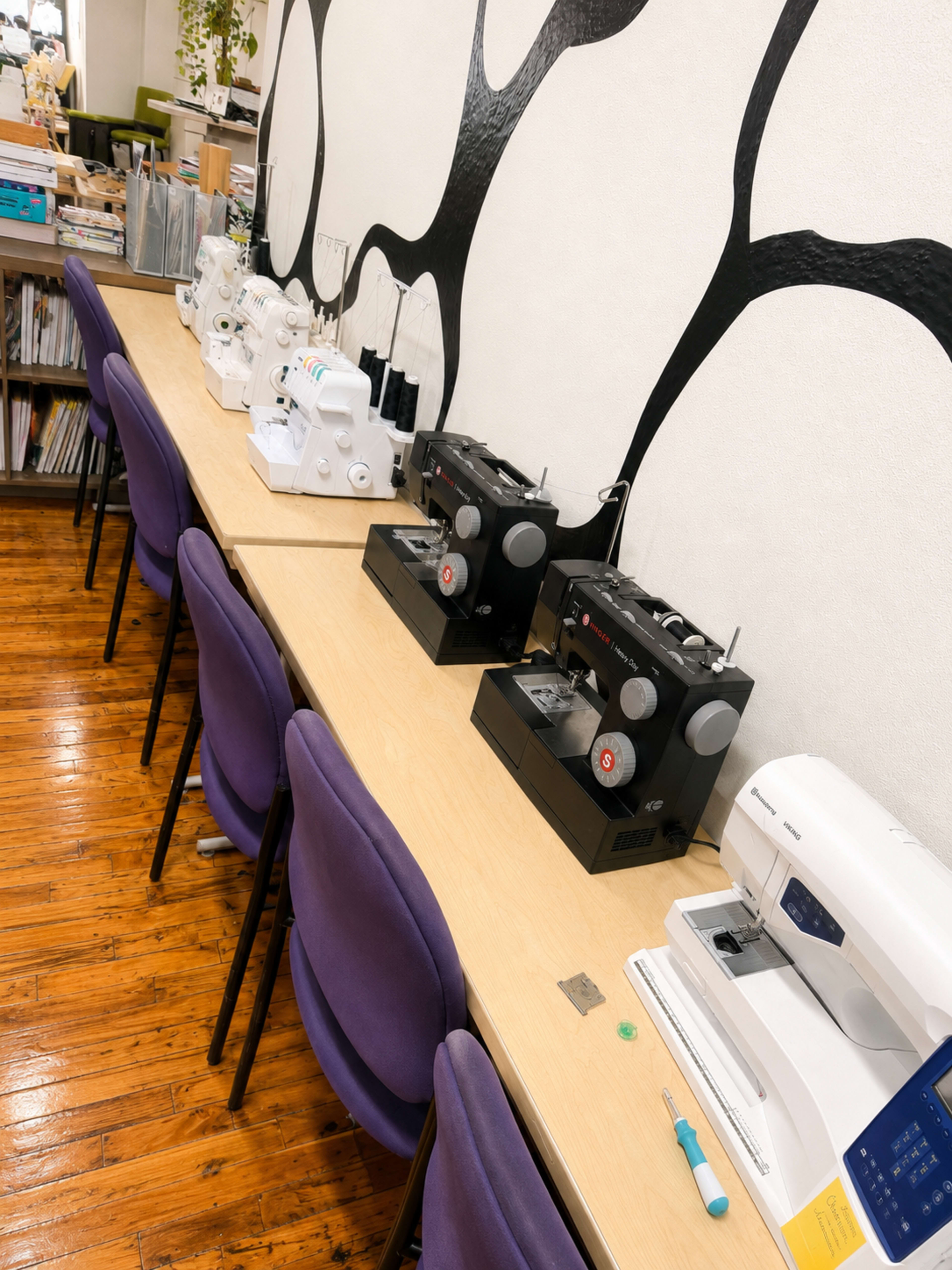 The image shows a row of sewing machines on a wooden table with purple chairs in a well-lit room.