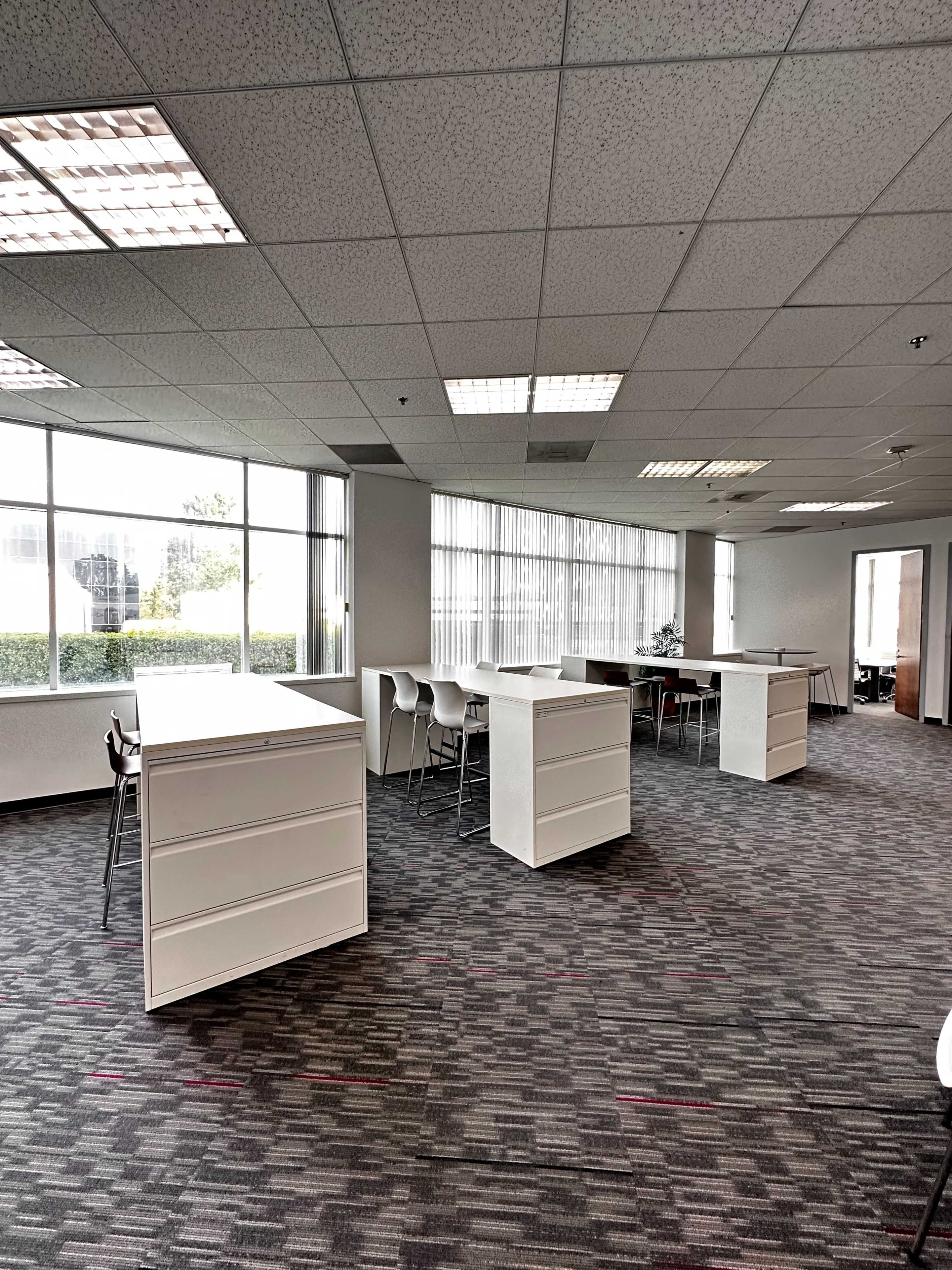 An office space with multiple white desks arranged in a carpeted area, illuminated by natural light from large windows.