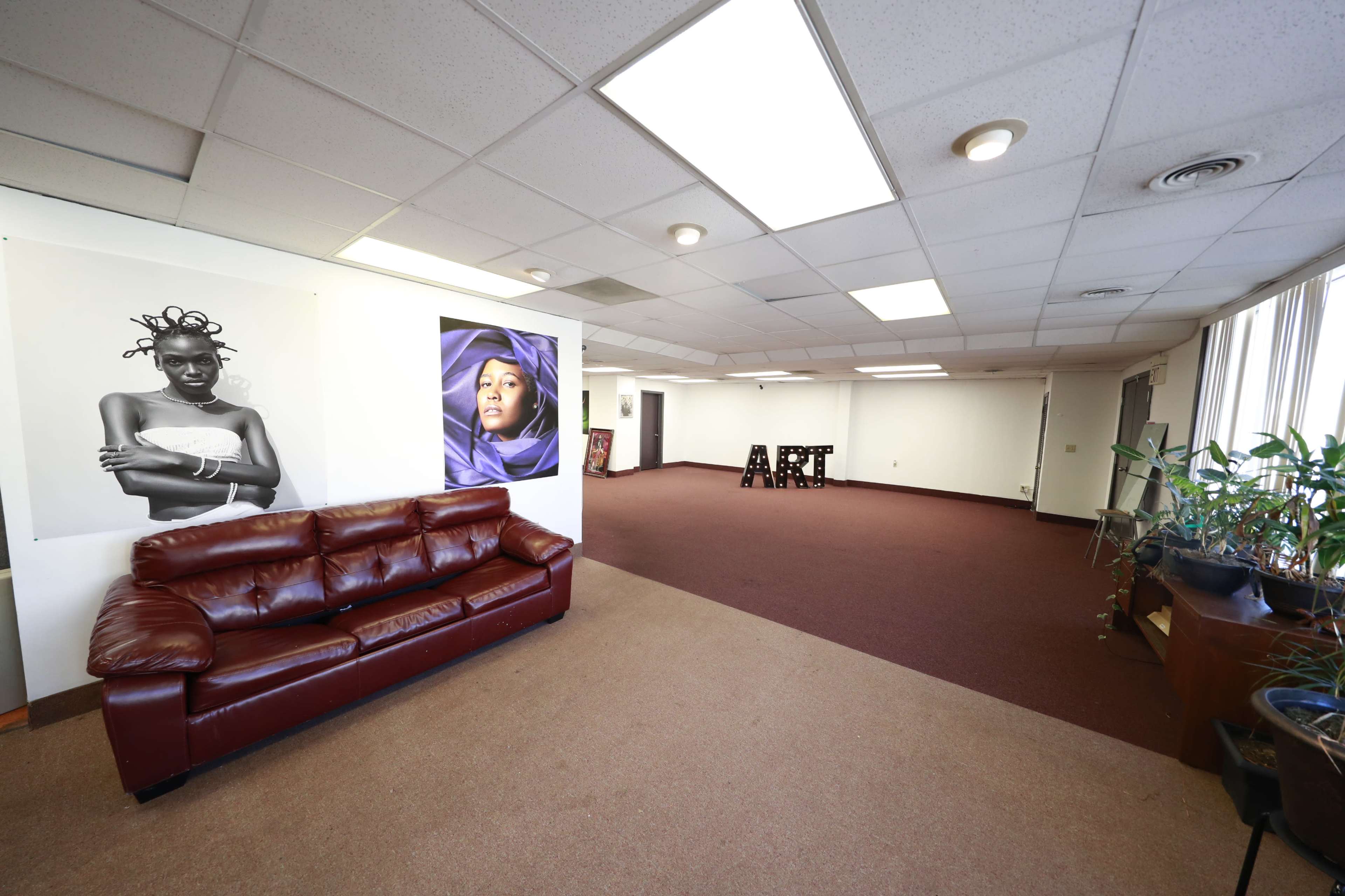 A spacious interior room with a brown leather sofa, large wall posters of women, and the word "ART" made of wooden letters on the floor.