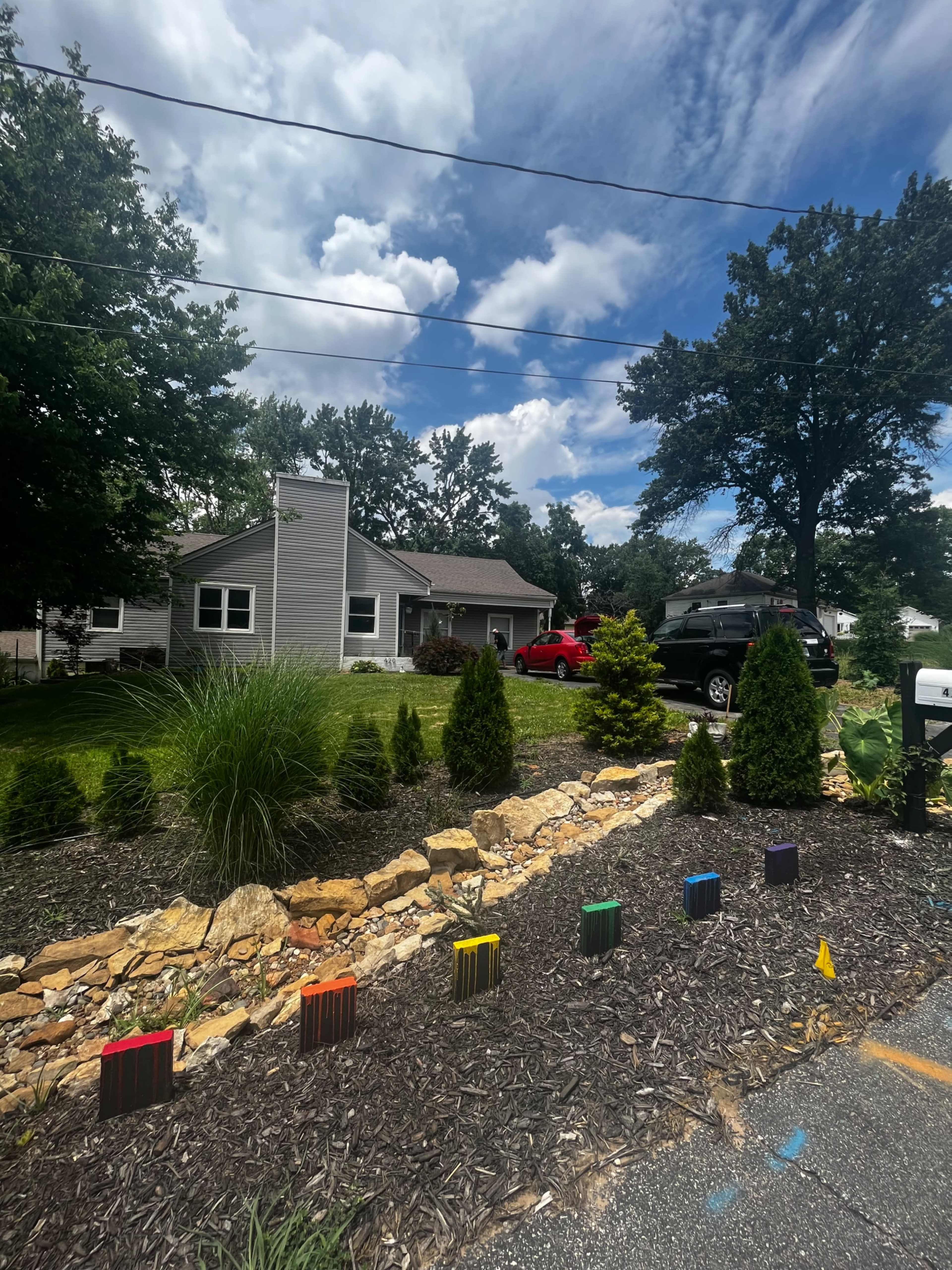 The image shows a suburban house with a well-maintained lawn, surrounded by decorative shrubs and colorful landscape edging, under a partly cloudy sky.