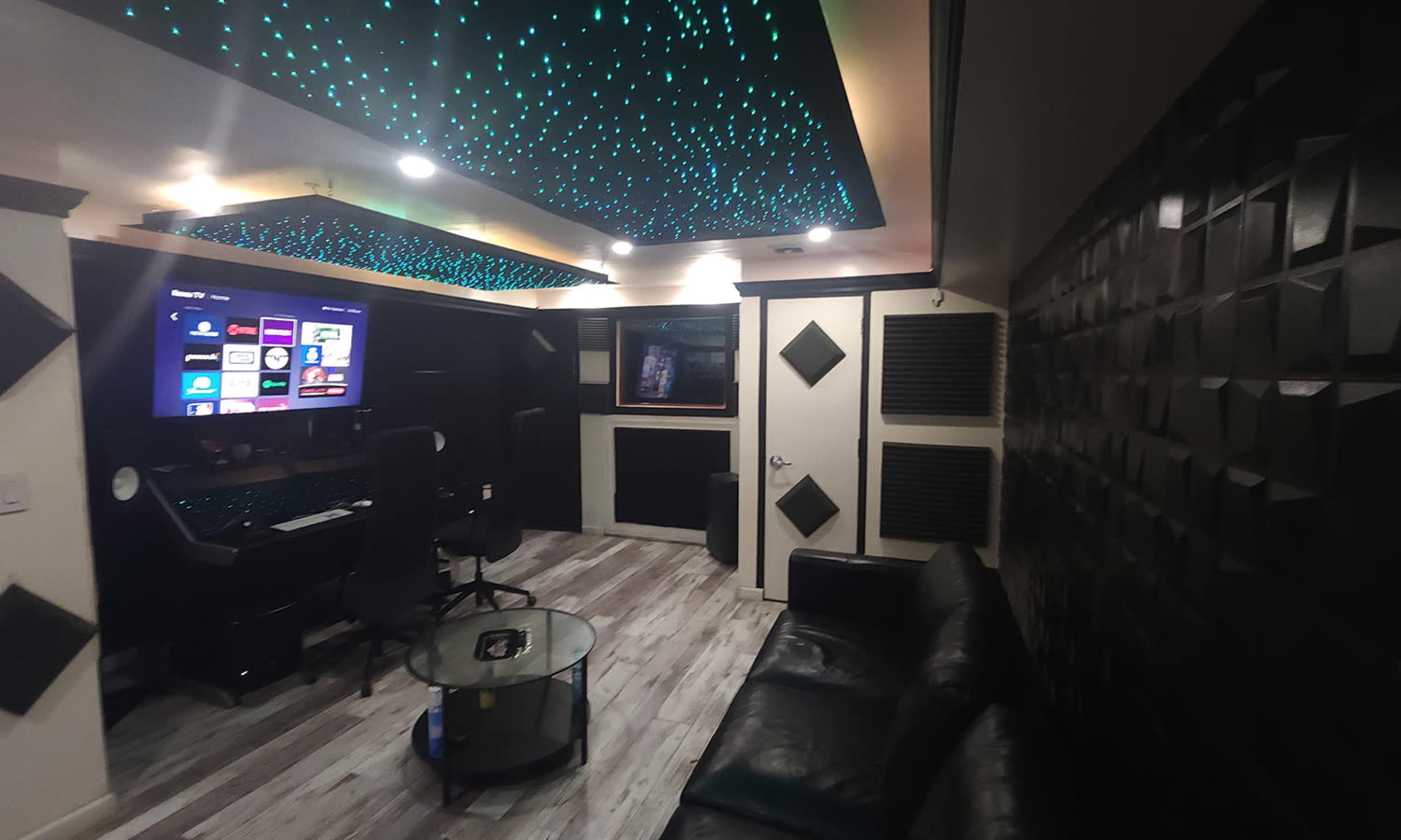 A modern recording studio with a black leather sofa, a glass coffee table, and a workstation equipped with a computer and large screen, illuminated by a starry ceiling.