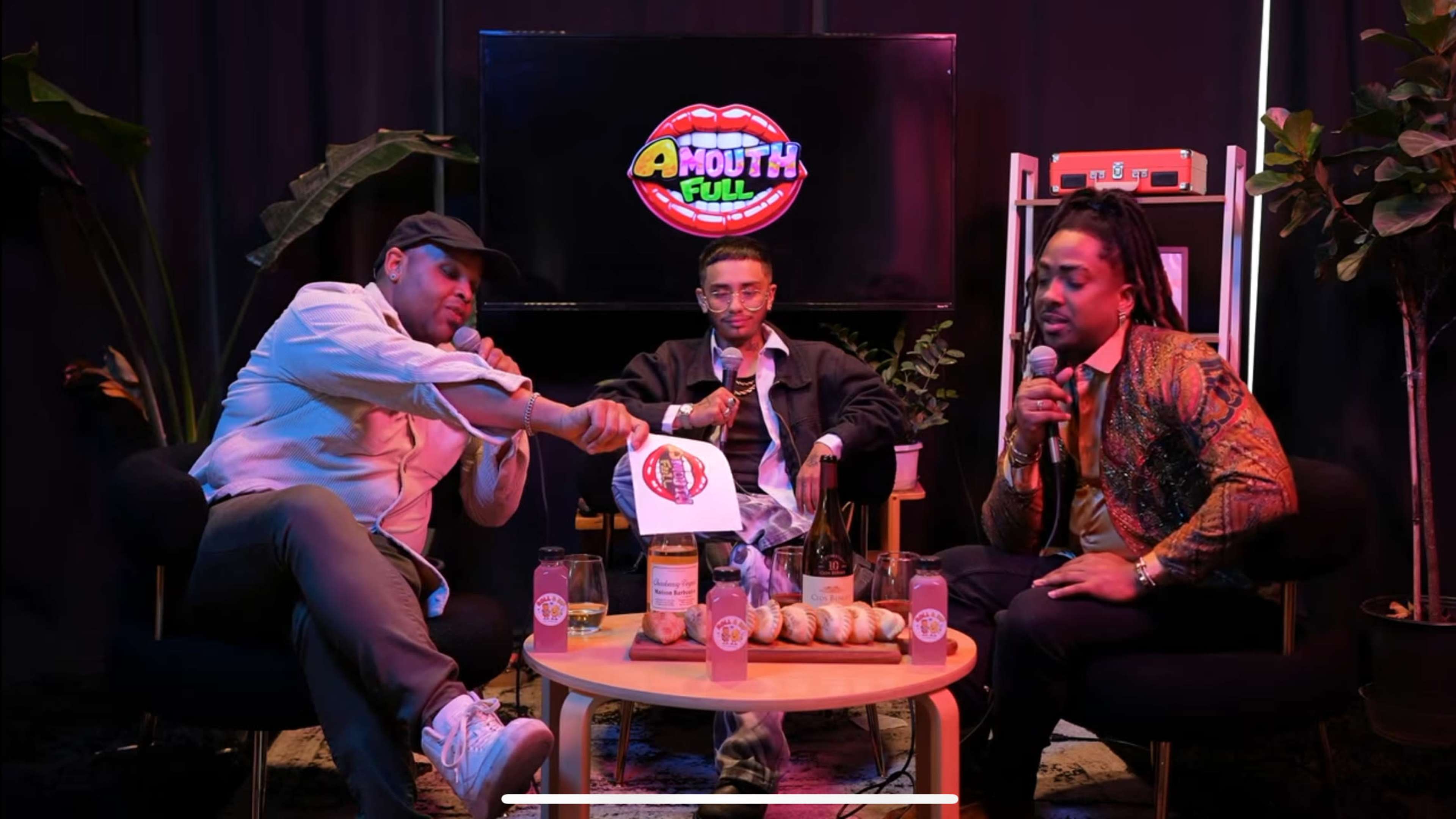 Three men sit on chairs around a coffee table holding microphones, with a colorful sign featuring the words "A Mouth Full" in the background.