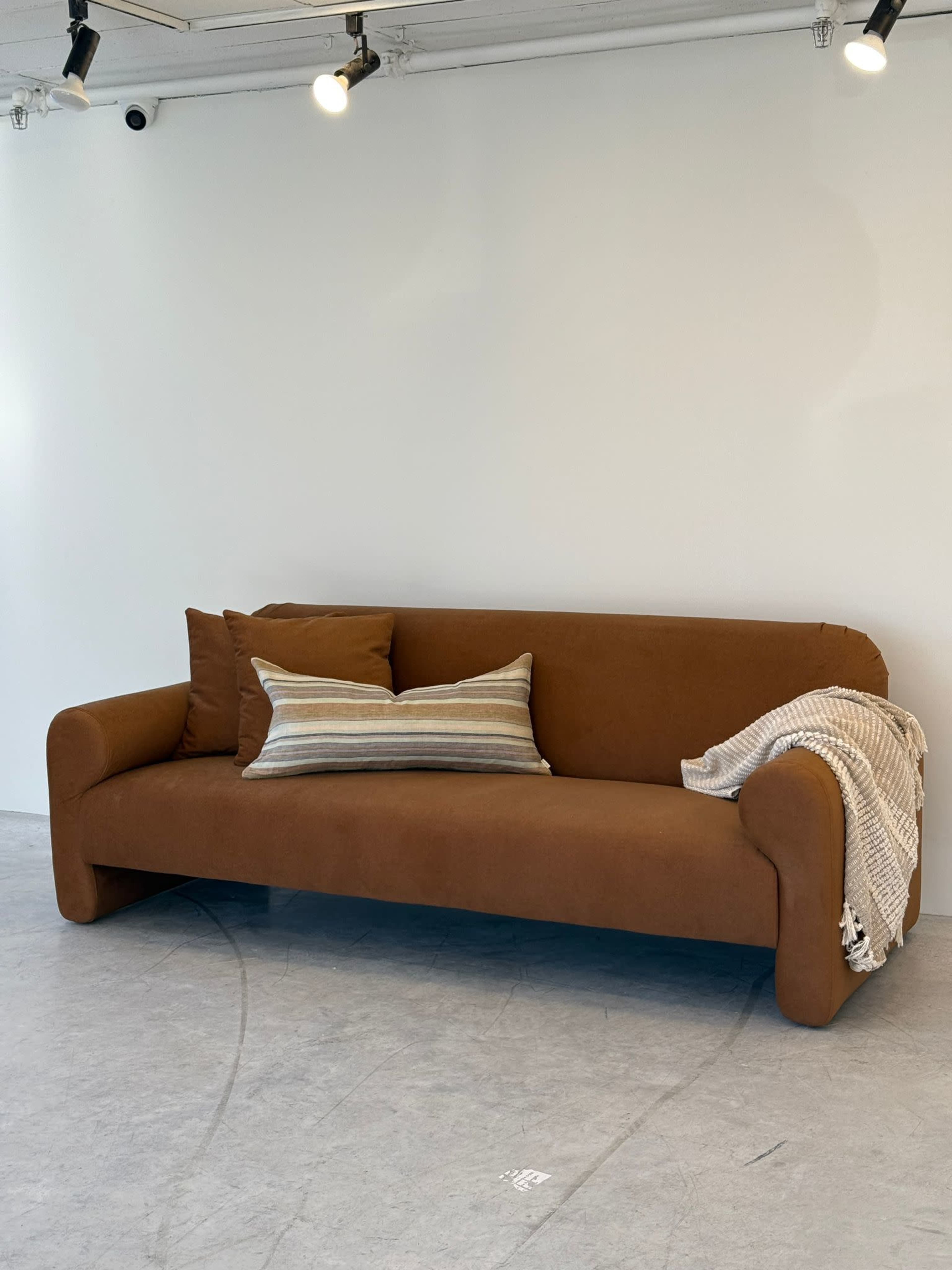 A brown sofa with two cushions and a striped throw blanket is positioned against a plain white wall in a well-lit room.