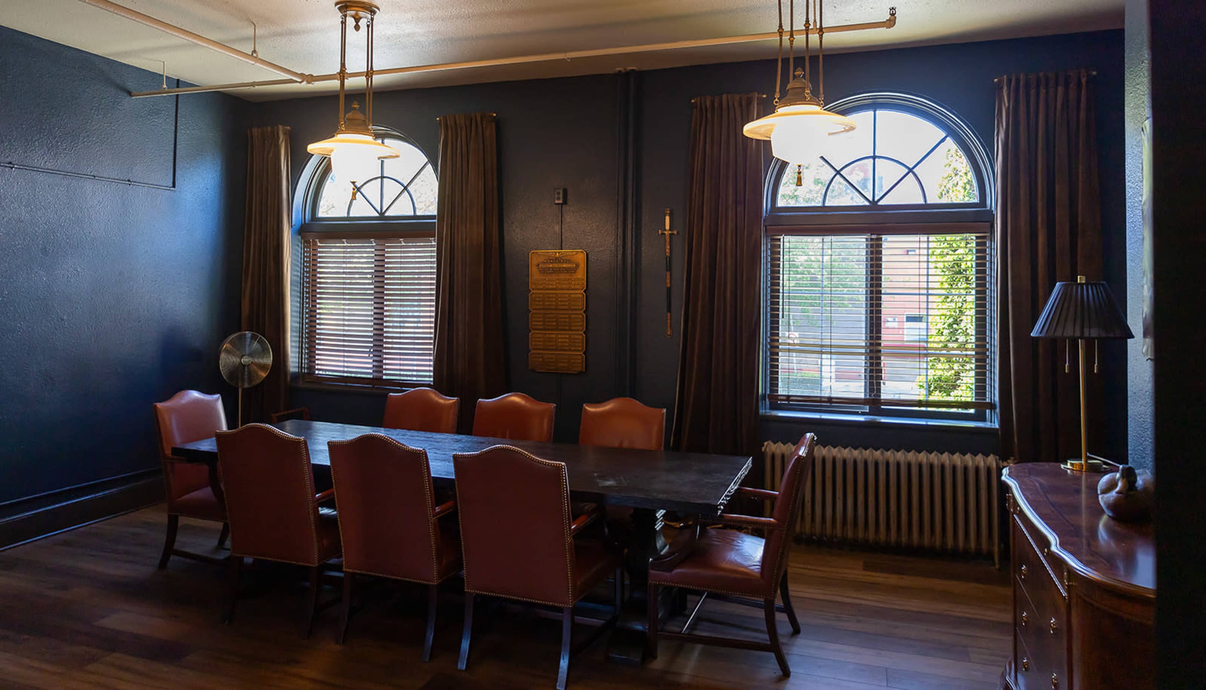The image shows a dining room with a long wooden table surrounded by eight upholstered chairs, two arched windows with dark curtains, and a radiator along the wall.