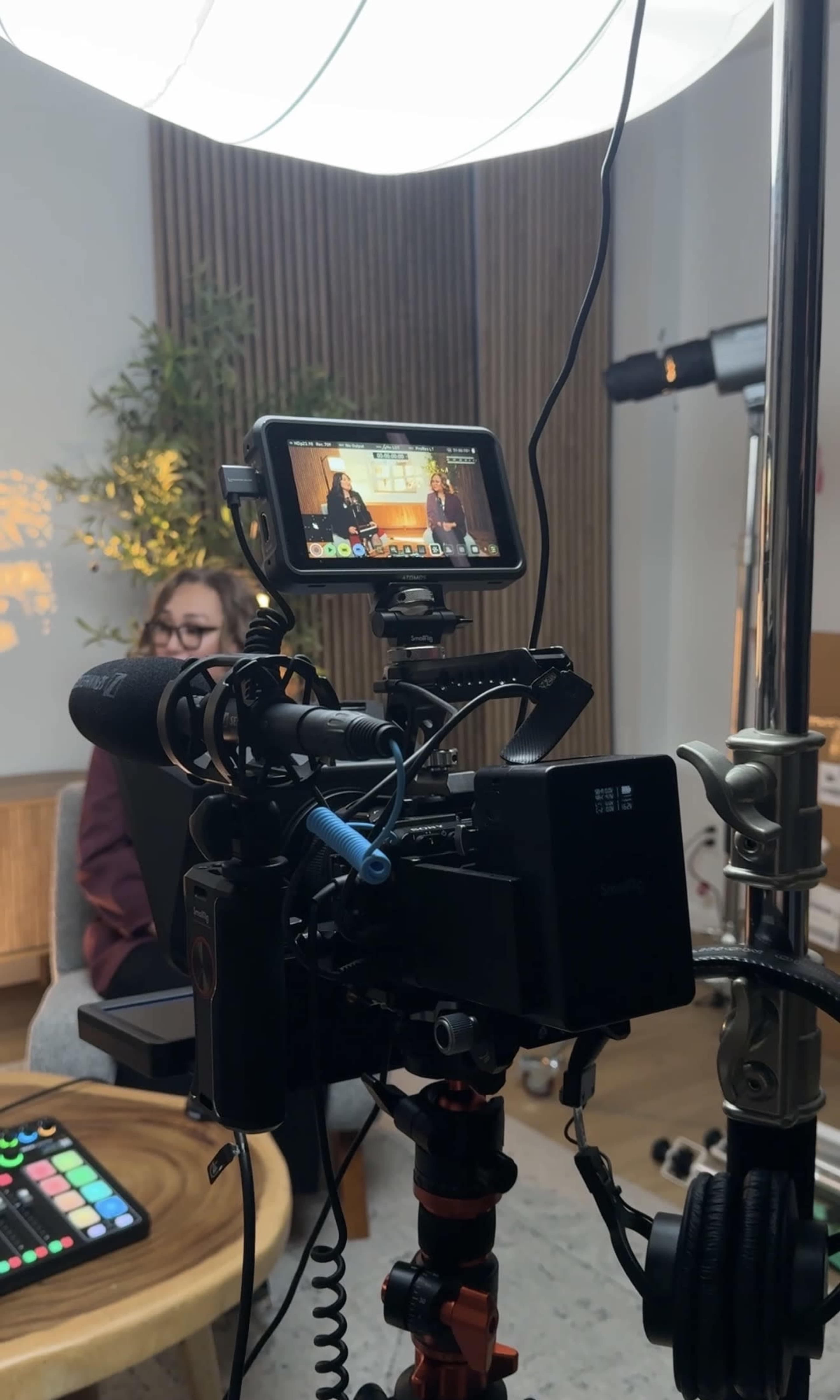 A camera setup on a tripod captures an interview scene, with a screen displaying two speakers in a well-lit studio environment.