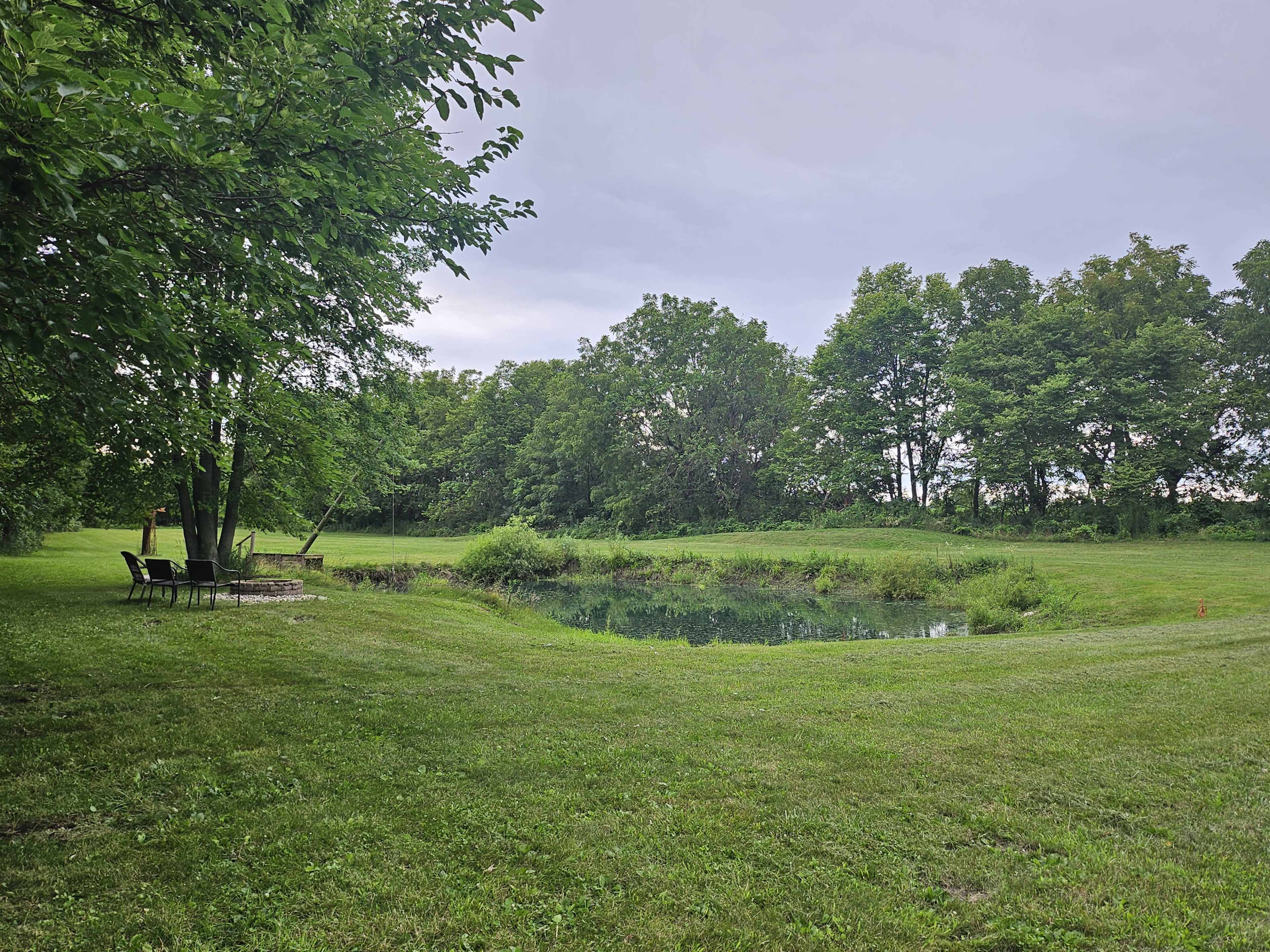 A tranquil pond is surrounded by grassy areas and trees, with a few chairs positioned nearby.