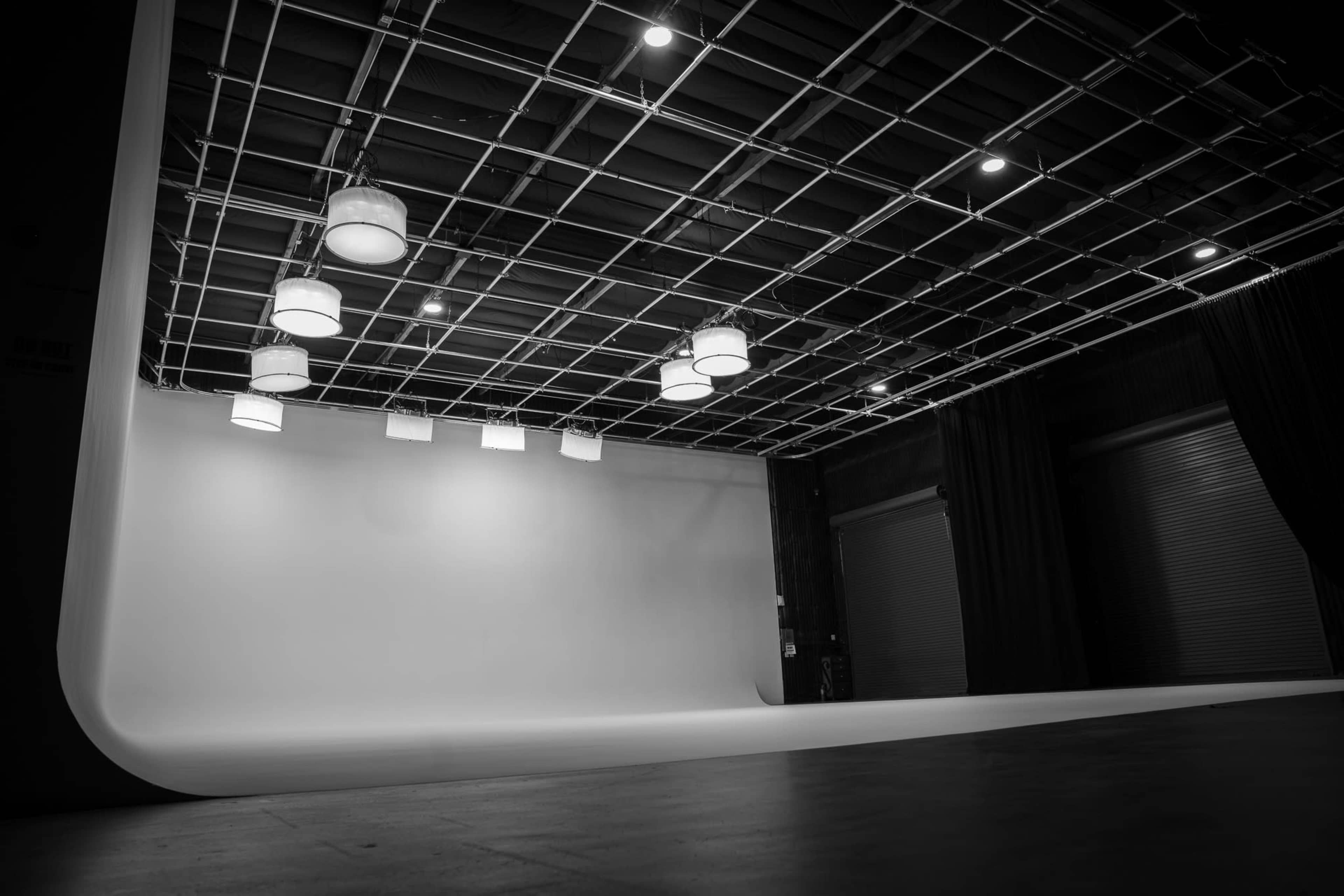The image shows an empty photography studio with a white backdrop and overhead lighting fixtures.