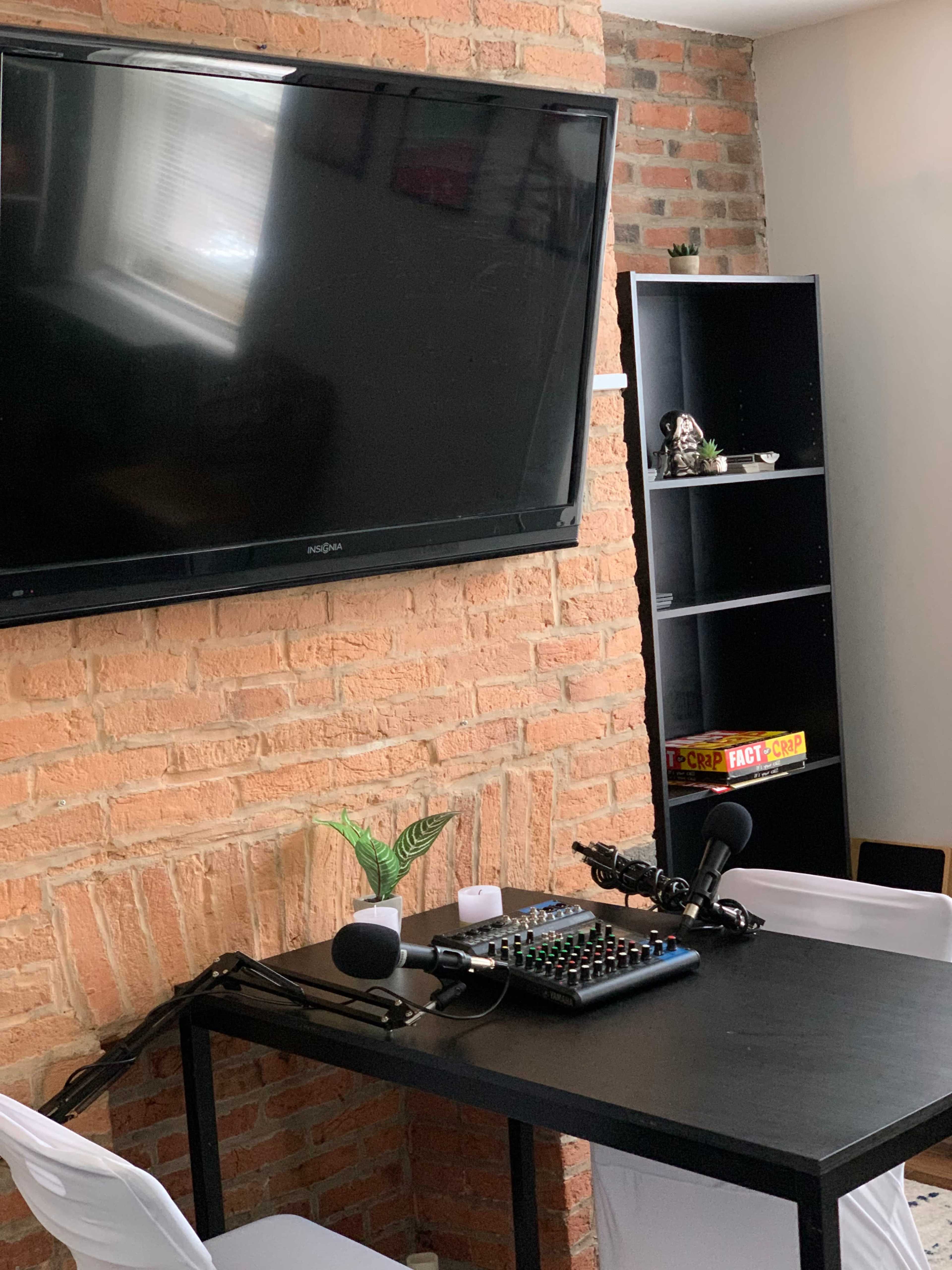 A room with a brick wall featuring a large TV mounted above a black desk equipped with microphones, a mixer, and two white chairs.