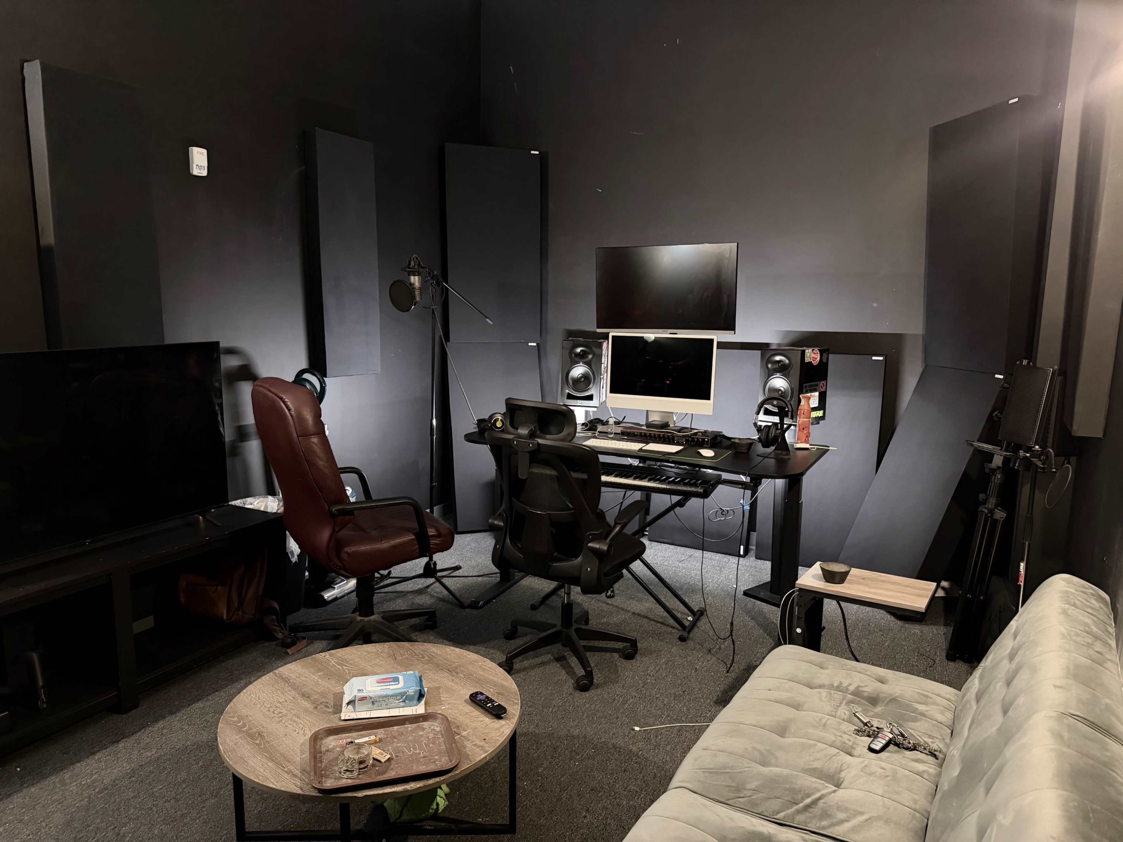 The image shows a dimly lit studio space featuring a desk with a computer and audio equipment, a comfortable chair, and a couch, surrounded by soundproofing panels.