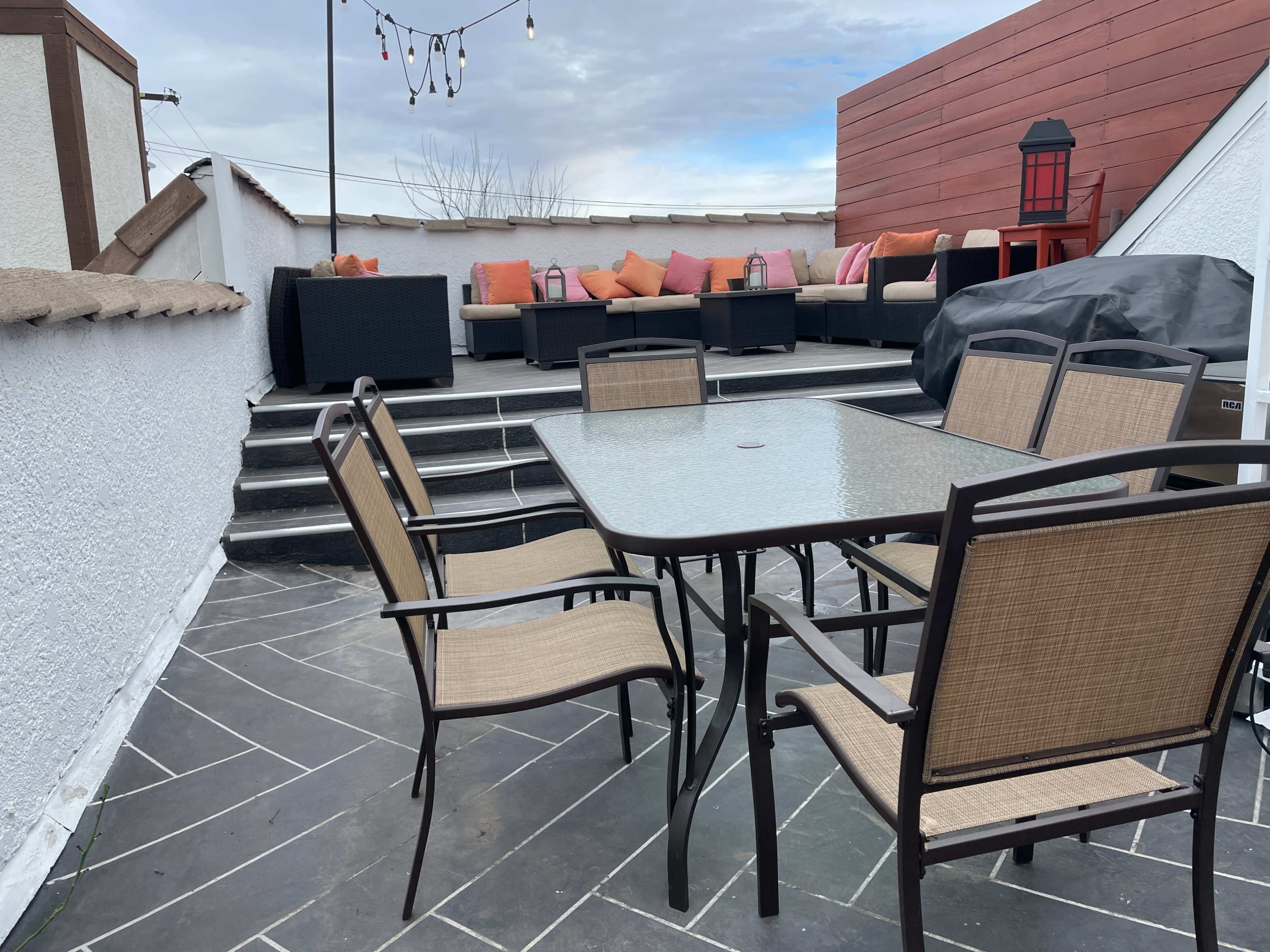 A rooftop terrace features a glass-top dining table surrounded by six chairs, with a seating area of black couches and colorful cushions in the background.