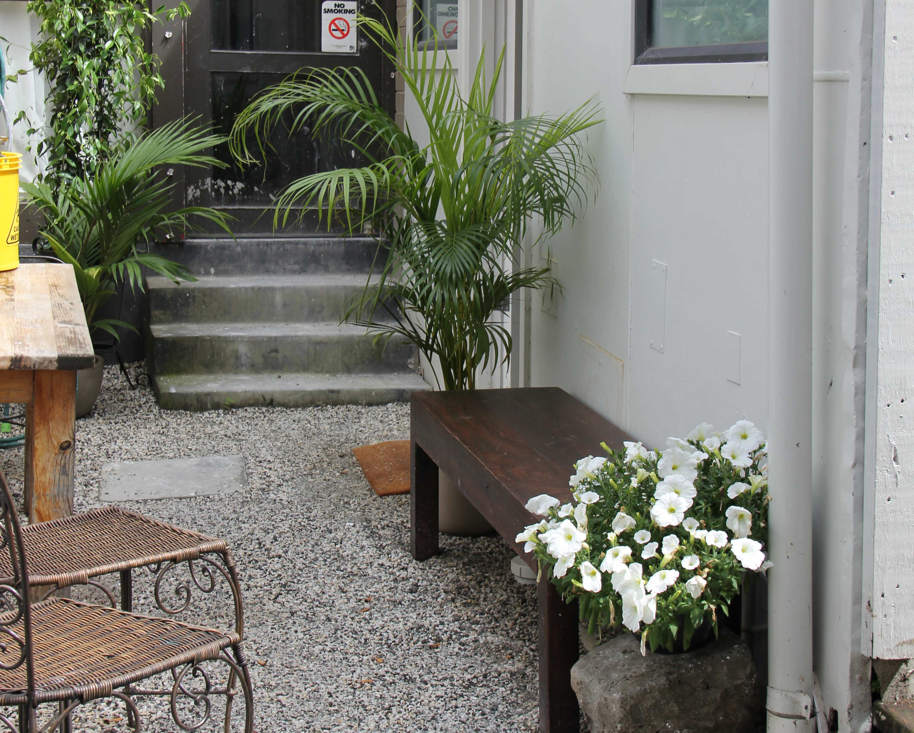 The image shows a narrow outdoor pathway with a wooden bench, a potted plant, and white flowers beside a staircase leading to a door.