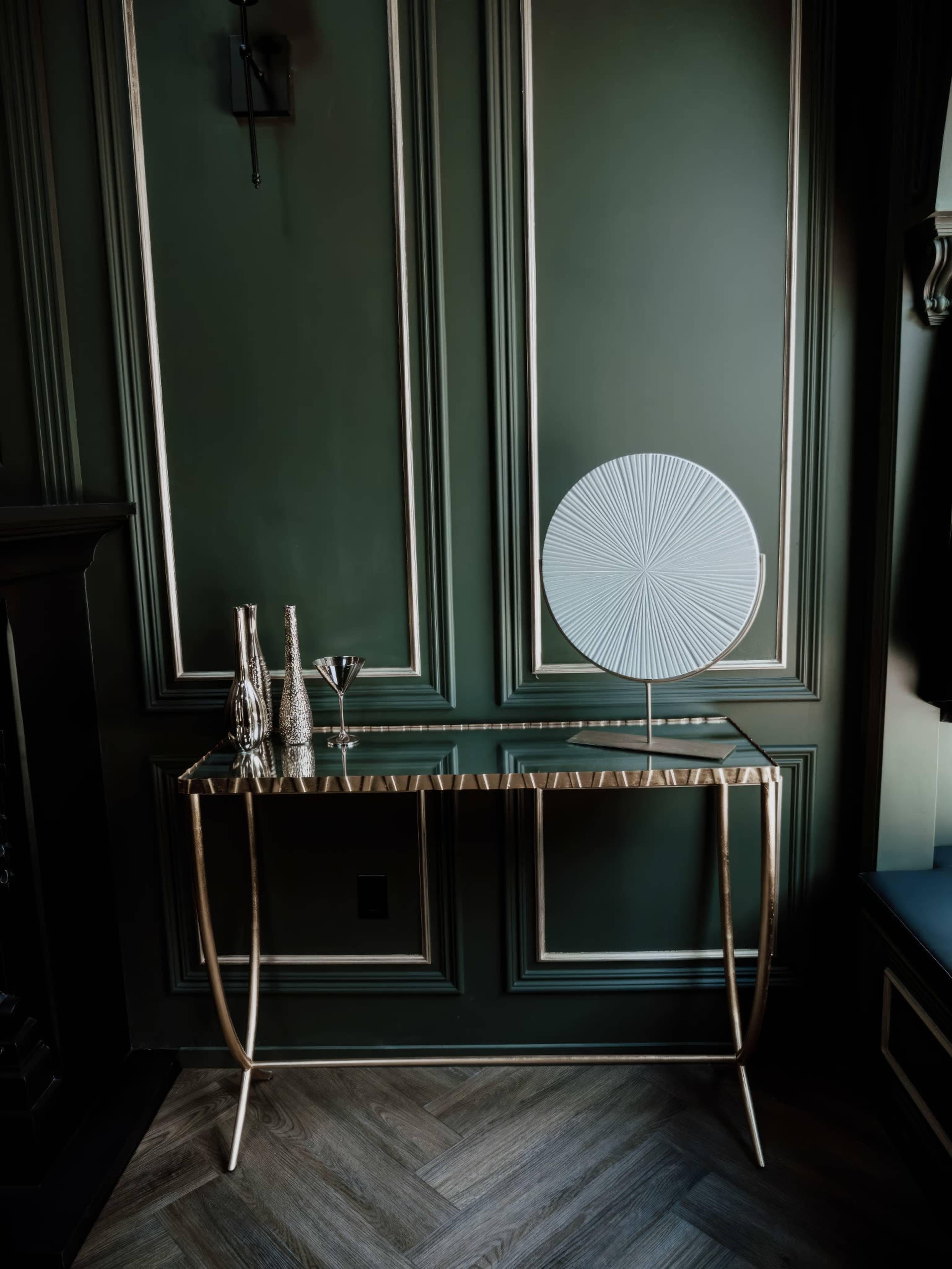 A glass-top console table with gold legs is positioned against a dark green wall, accompanied by three silver vases and a decorative circular plate.