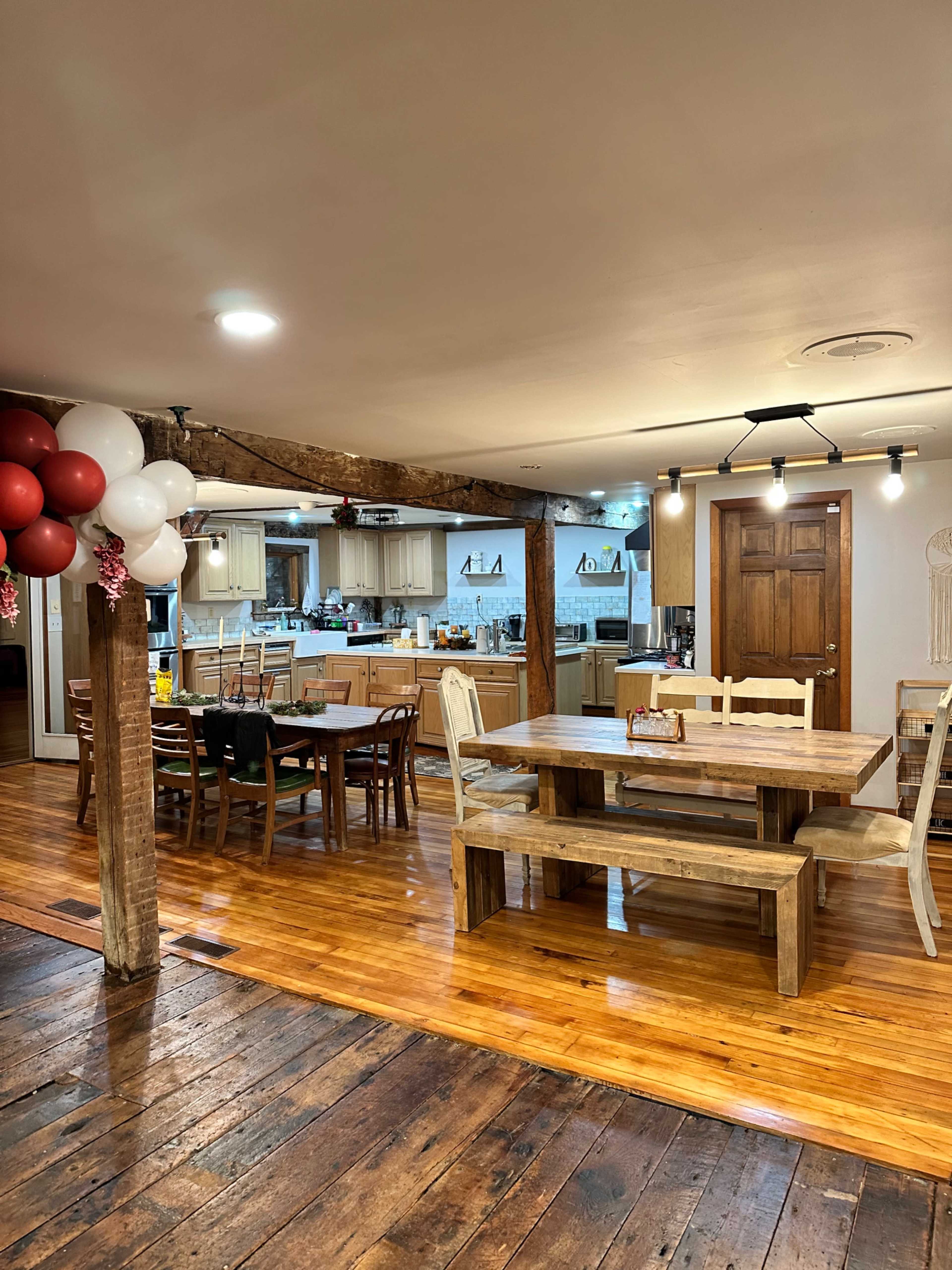 The image shows a spacious kitchen and dining area with wooden flooring, a large wooden dining table, several chairs, and red and white balloons near a wooden support beam.