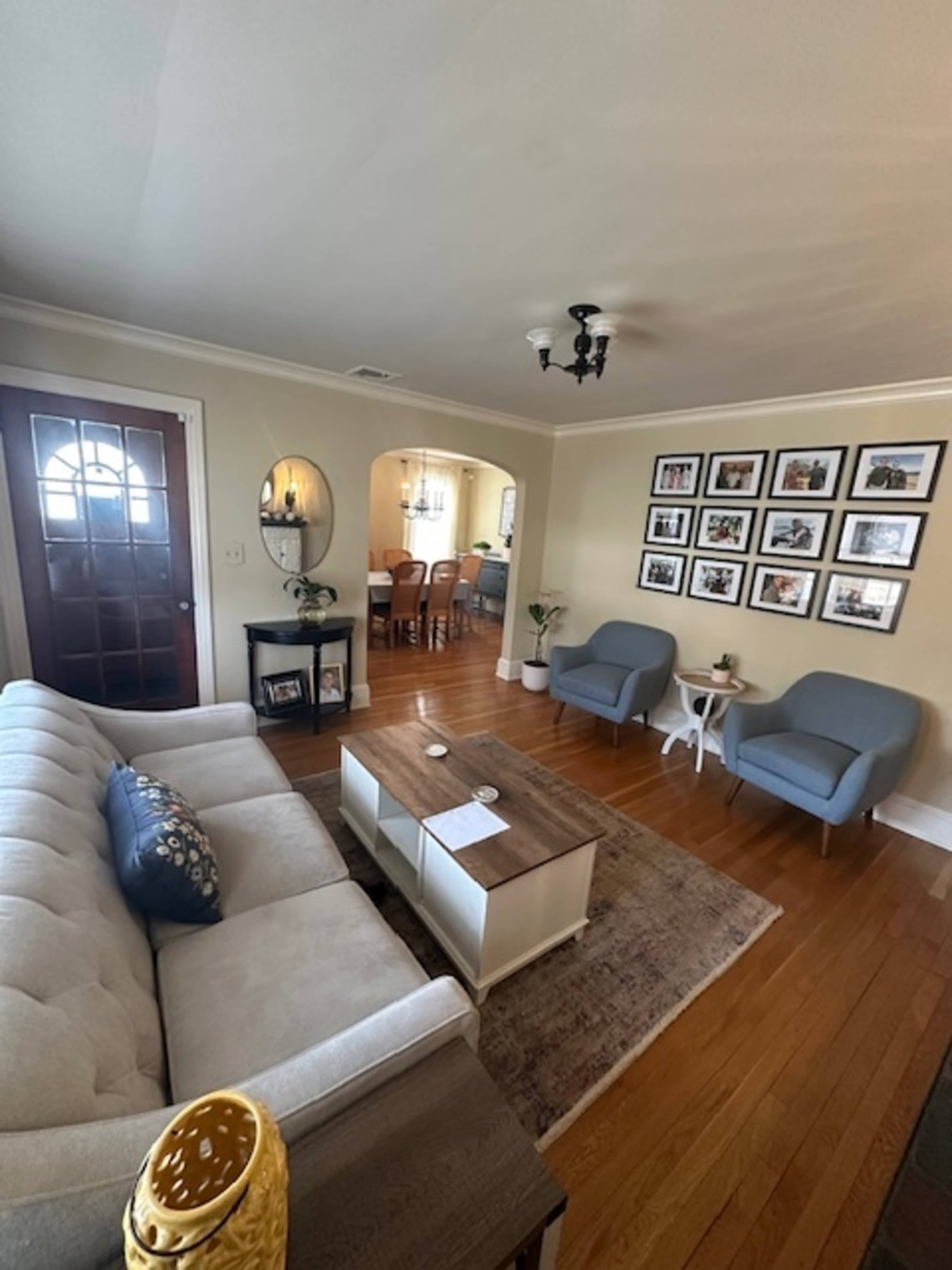 The image shows a living room with a light-colored sofa, two blue chairs, a wooden coffee table, and a decorative wall filled with framed photographs, leading to a dining area in the background.