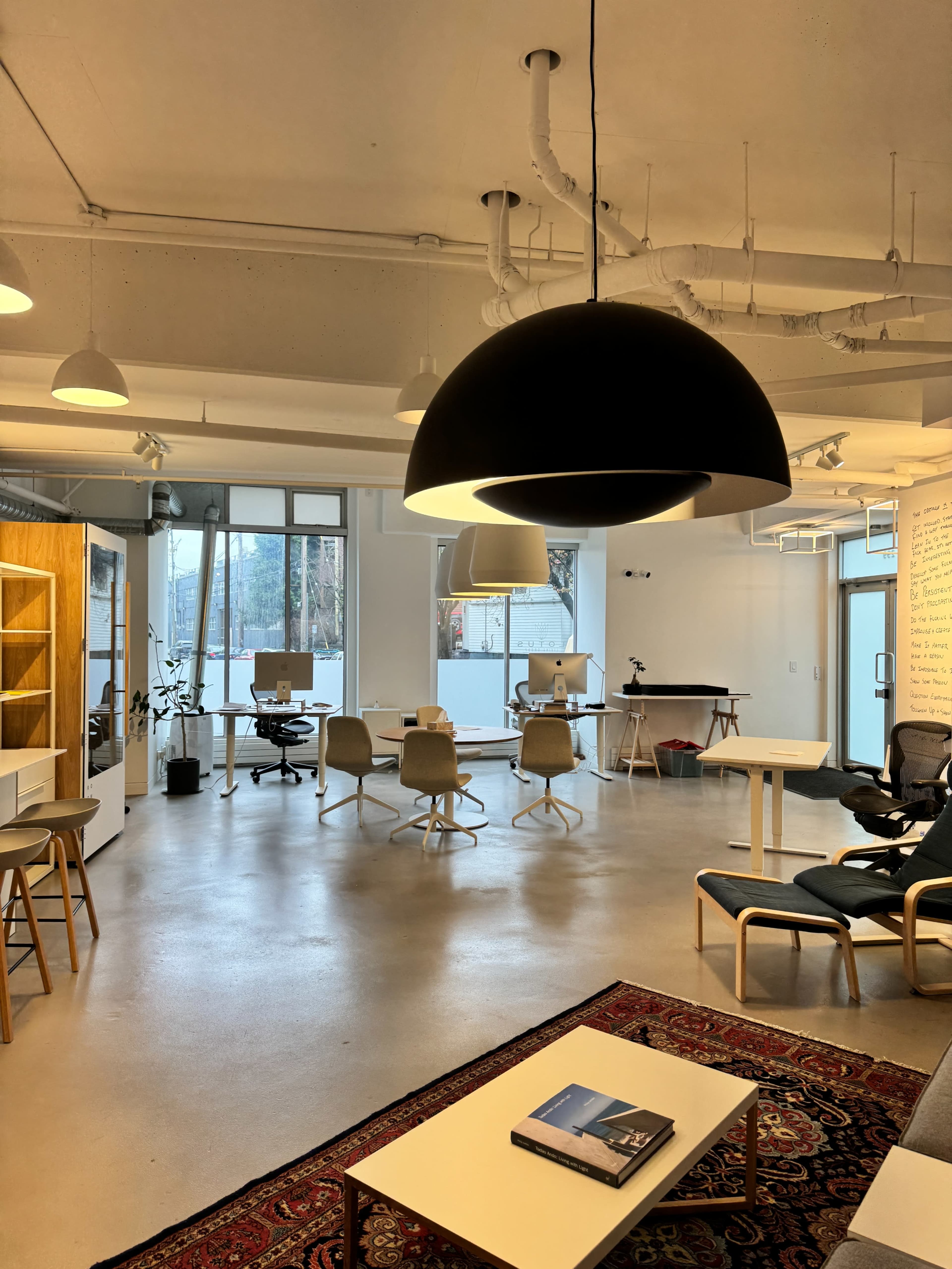 A modern office space with white walls, large windows, a black hanging light fixture, desks with computers, and a seating area featuring a rug and a coffee table.