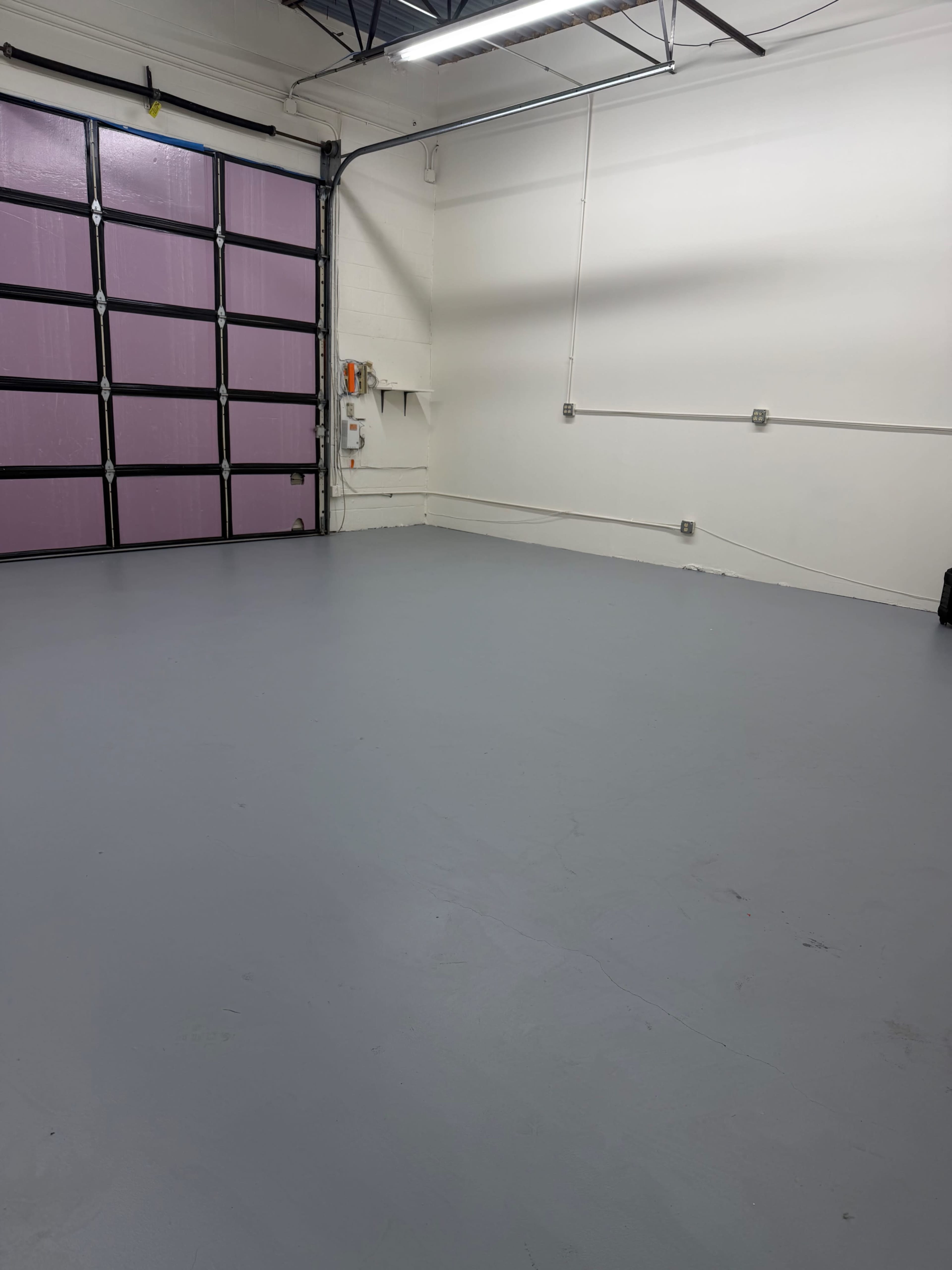 The image shows a large, empty room with gray flooring and a pink garage door on one side.