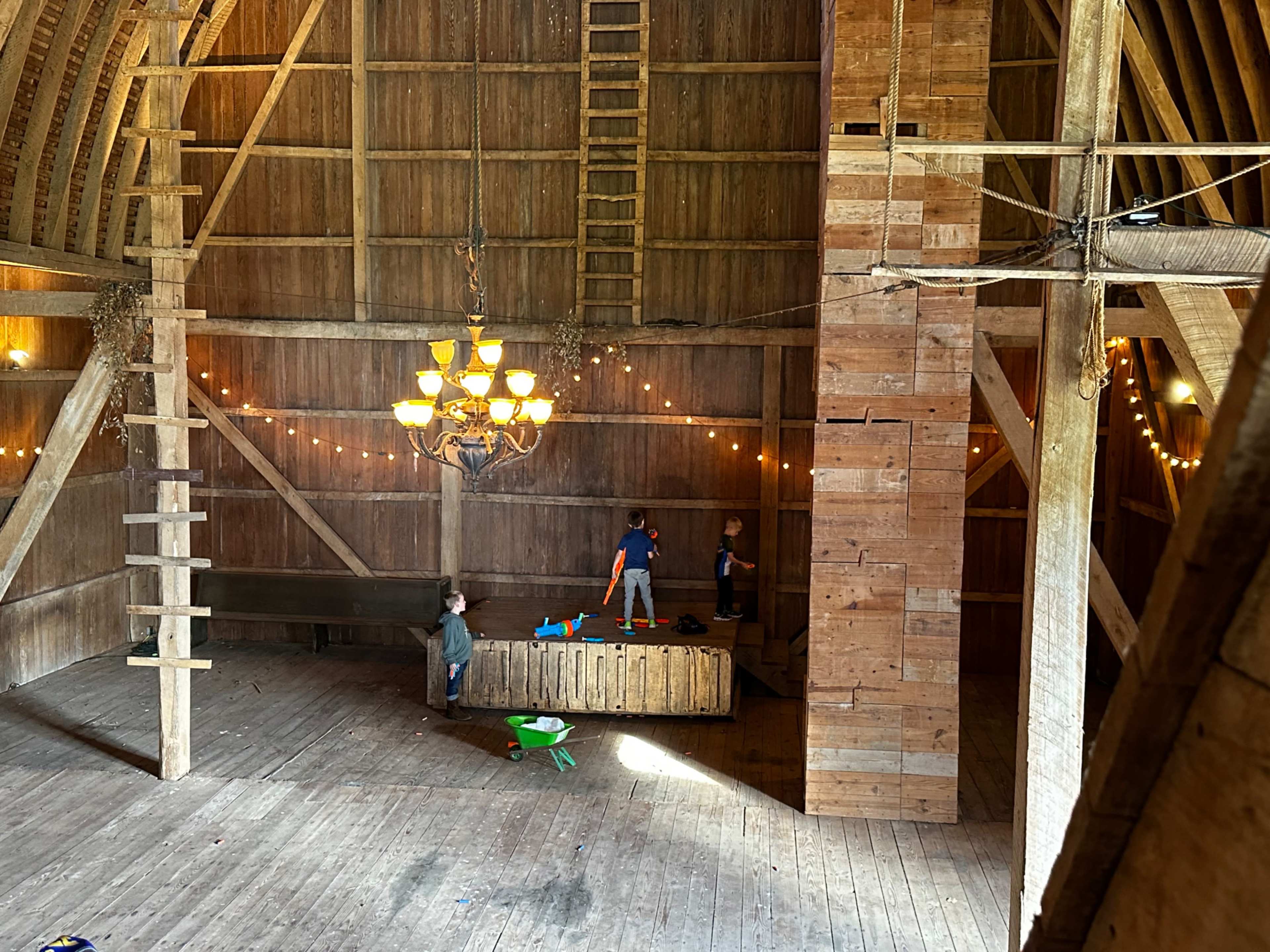 Three children play inside a spacious, wooden barn illuminated by a chandelier and string lights.