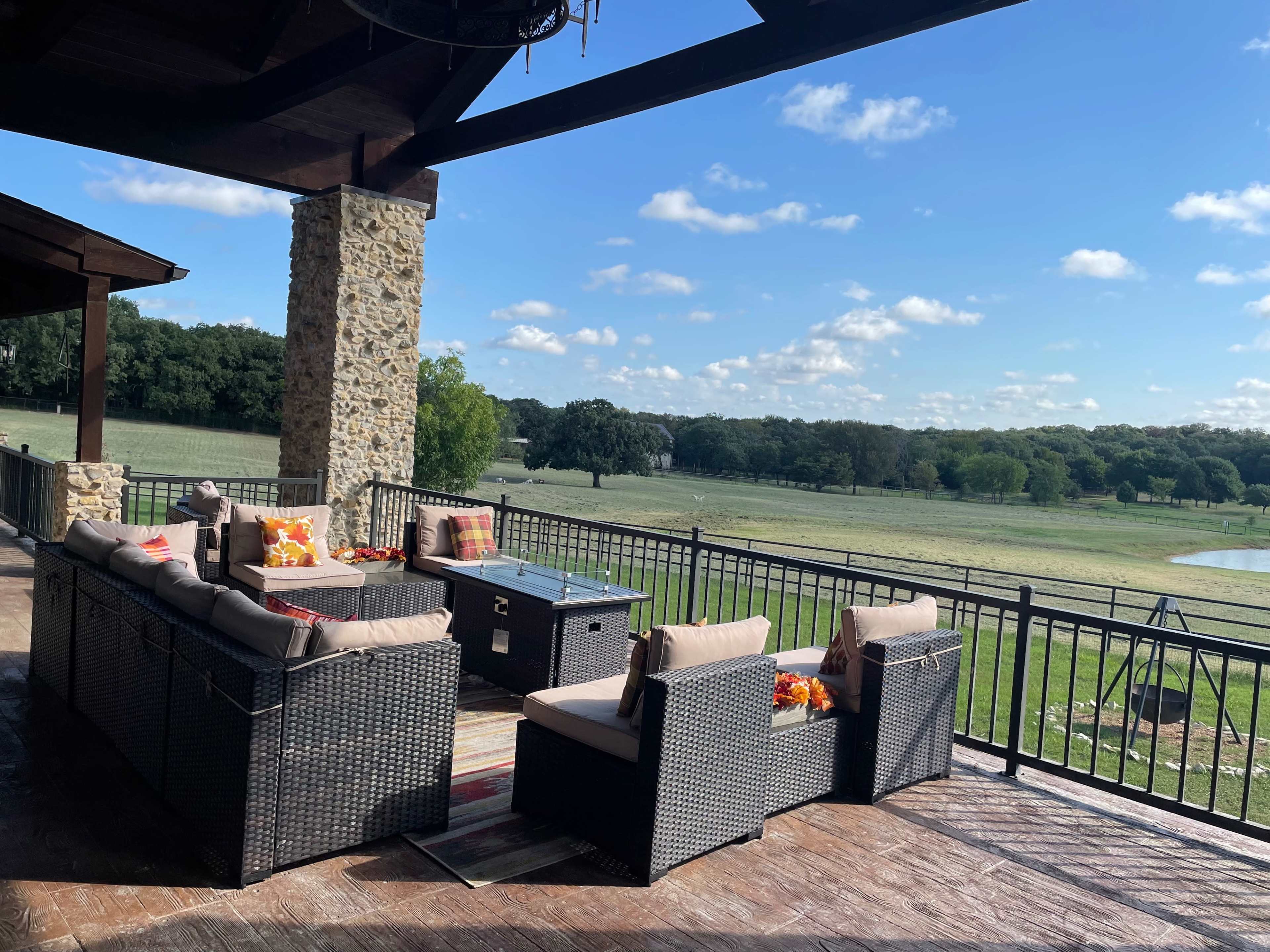 A spacious patio features wicker furniture, a coffee table, and a view of a grassy landscape with trees under a partly cloudy sky.
