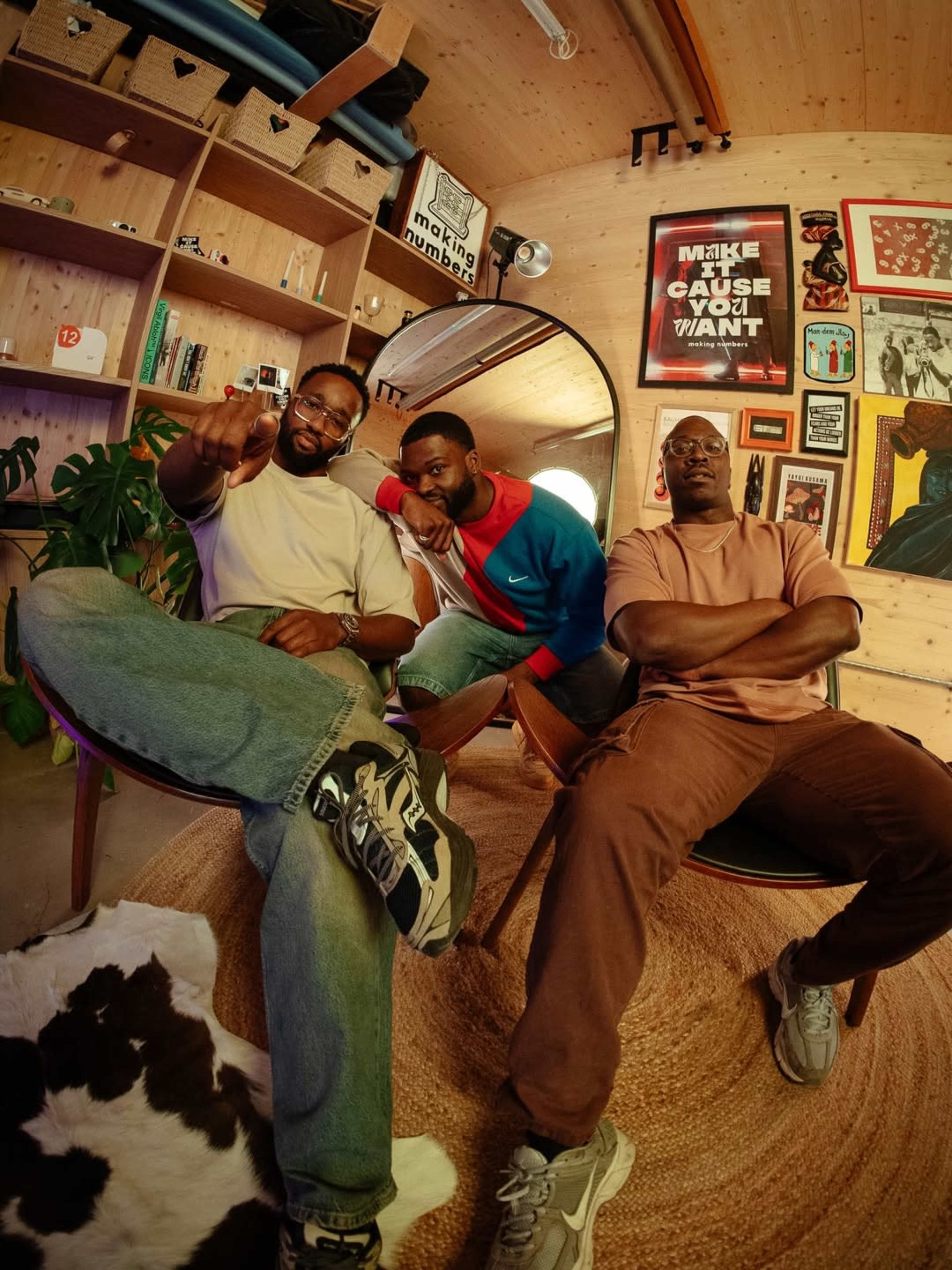 Three men pose for a photo in a warmly lit room filled with wooden shelves, artwork, and a decorative rug.