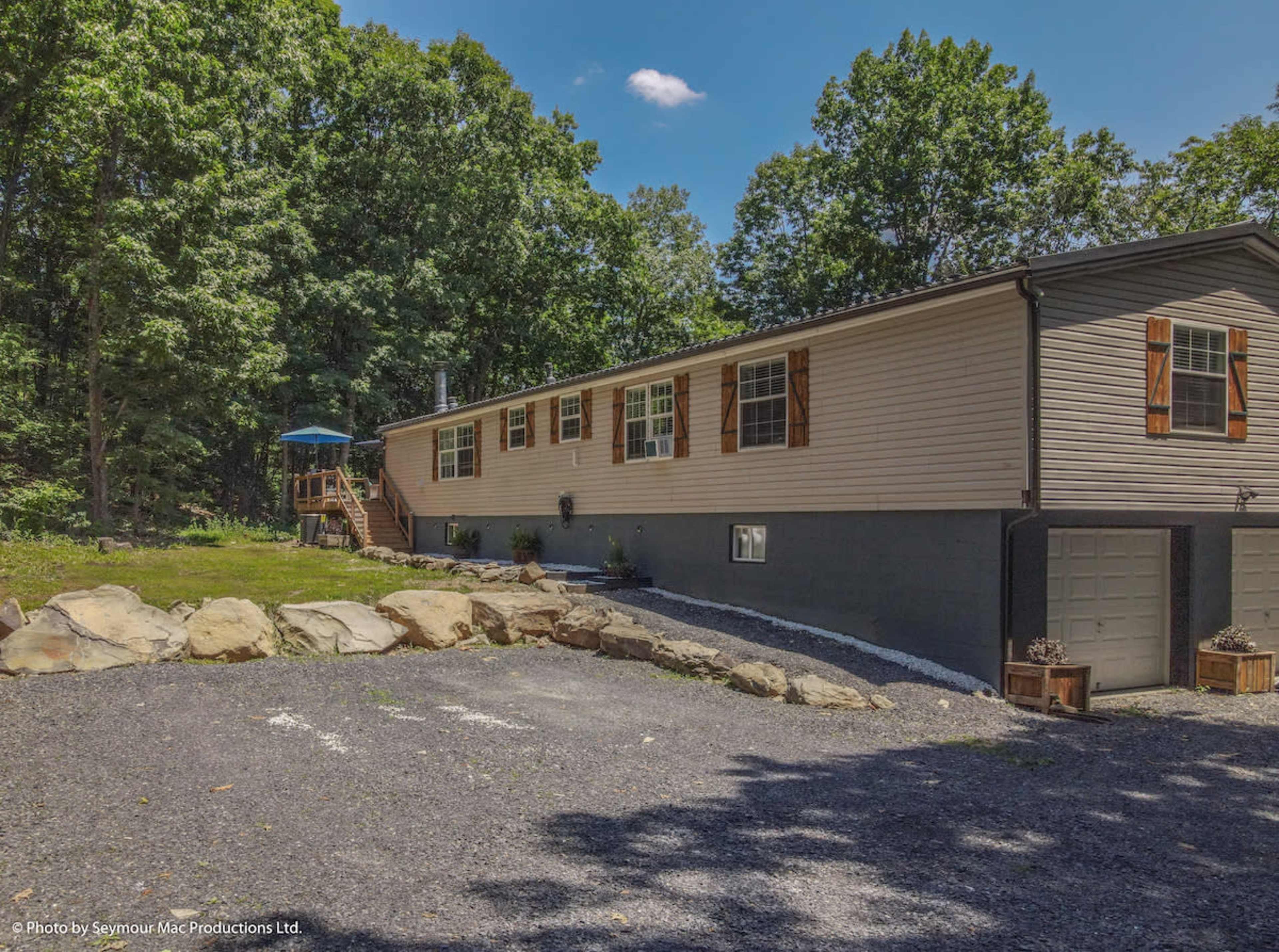 A large, single-story home with a gravel driveway is set against a backdrop of trees, featuring a porch and stone landscaping.