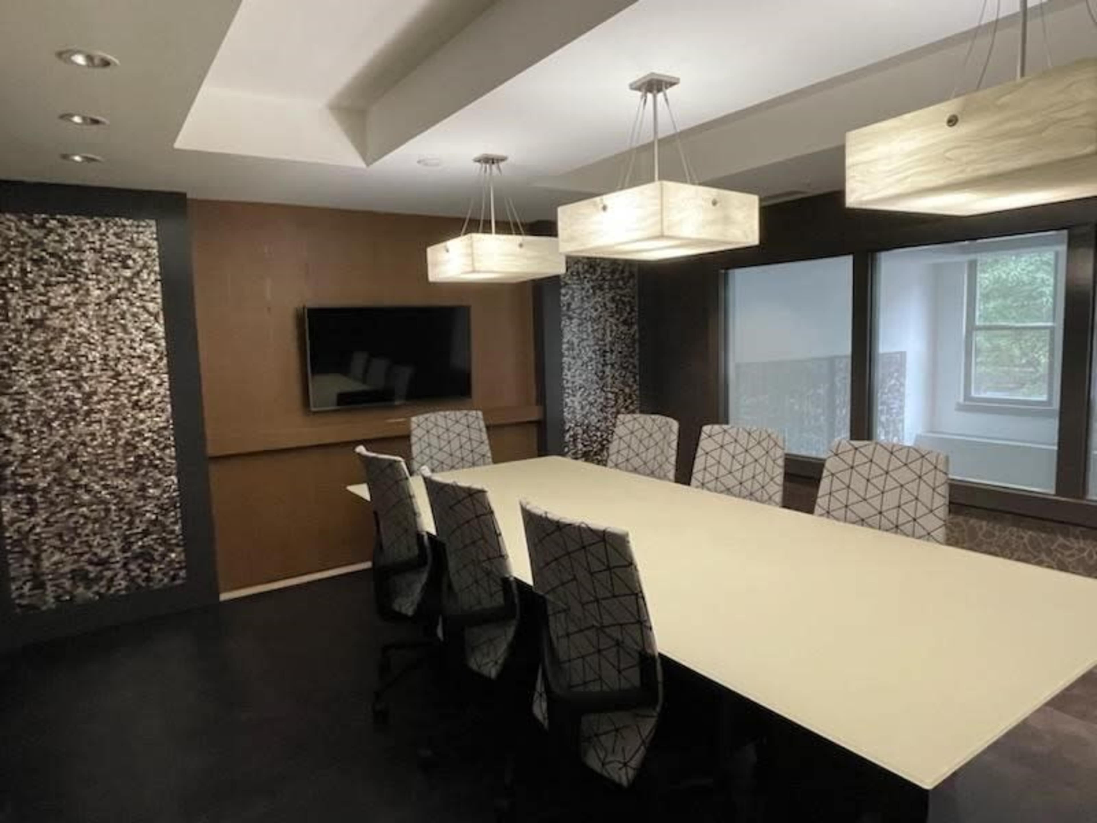 A modern conference room with a long table, eight patterned chairs, a wall-mounted television, and pendant lighting.
