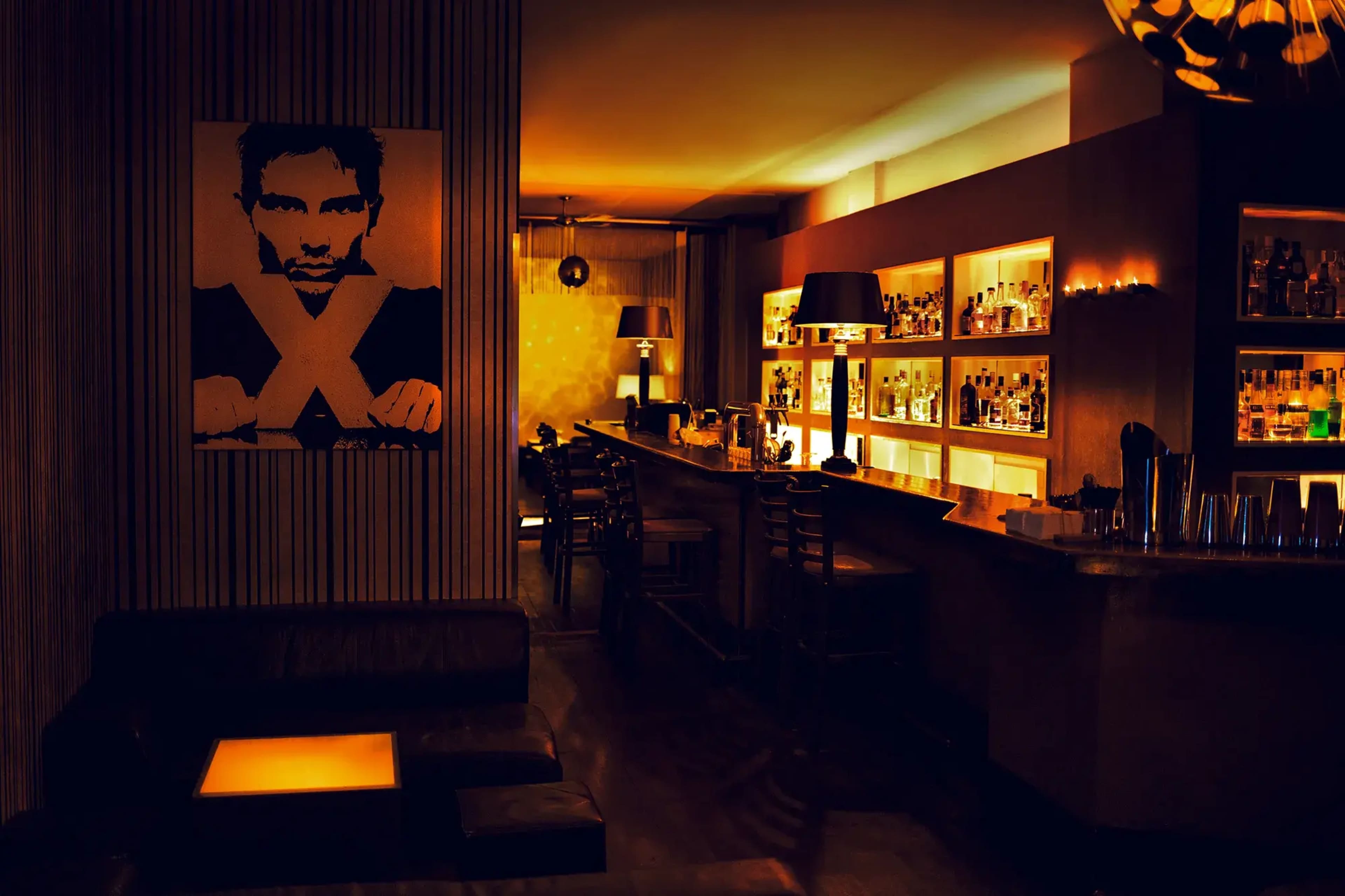 A dimly lit bar features a prominent artwork of a man on the wall, with shelves filled with bottles behind the counter.