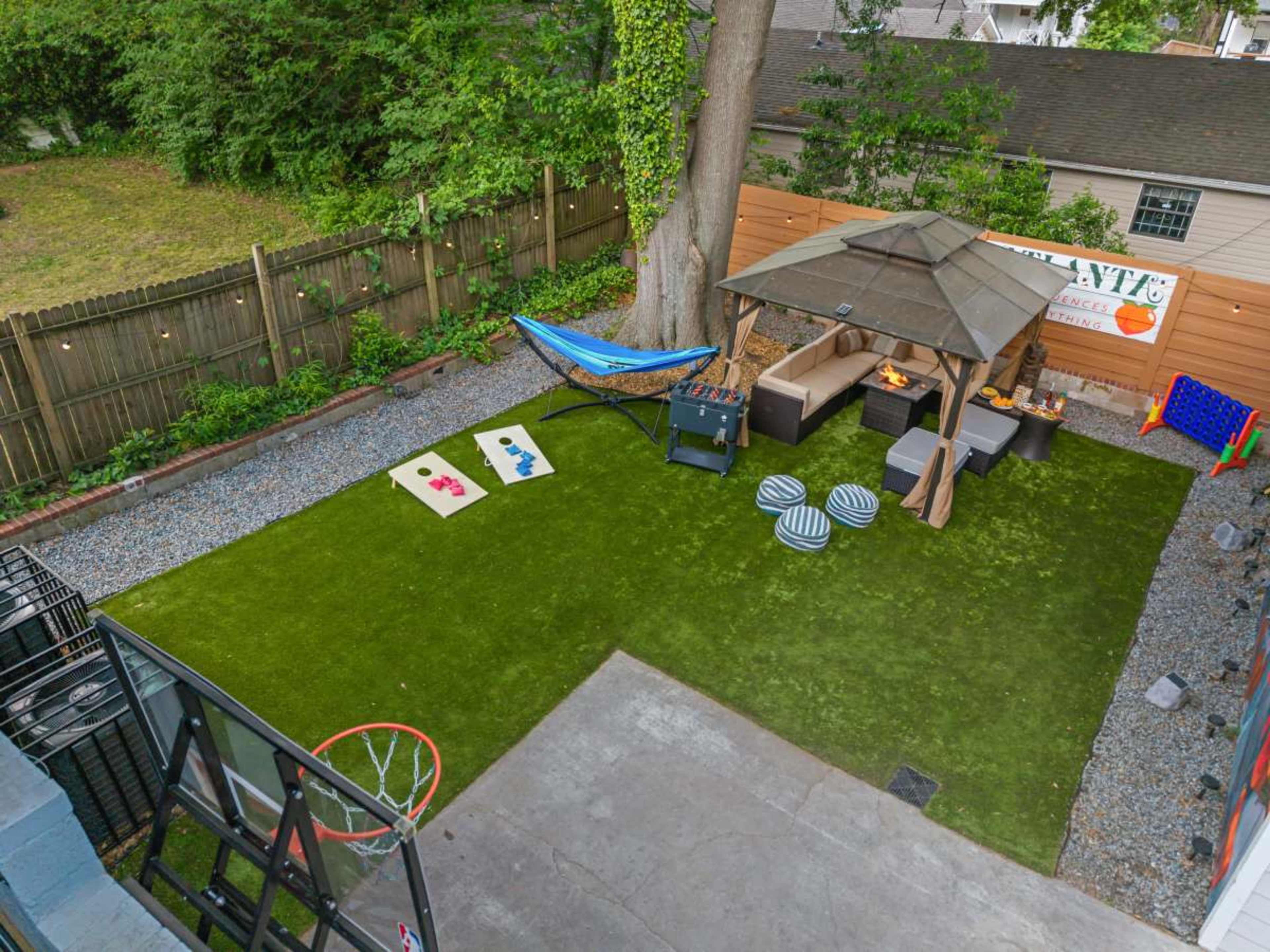 The image shows a well-designed backyard with a patio, a shaded seating area under a gazebo, a hammock, and games like cornhole and basketball.
