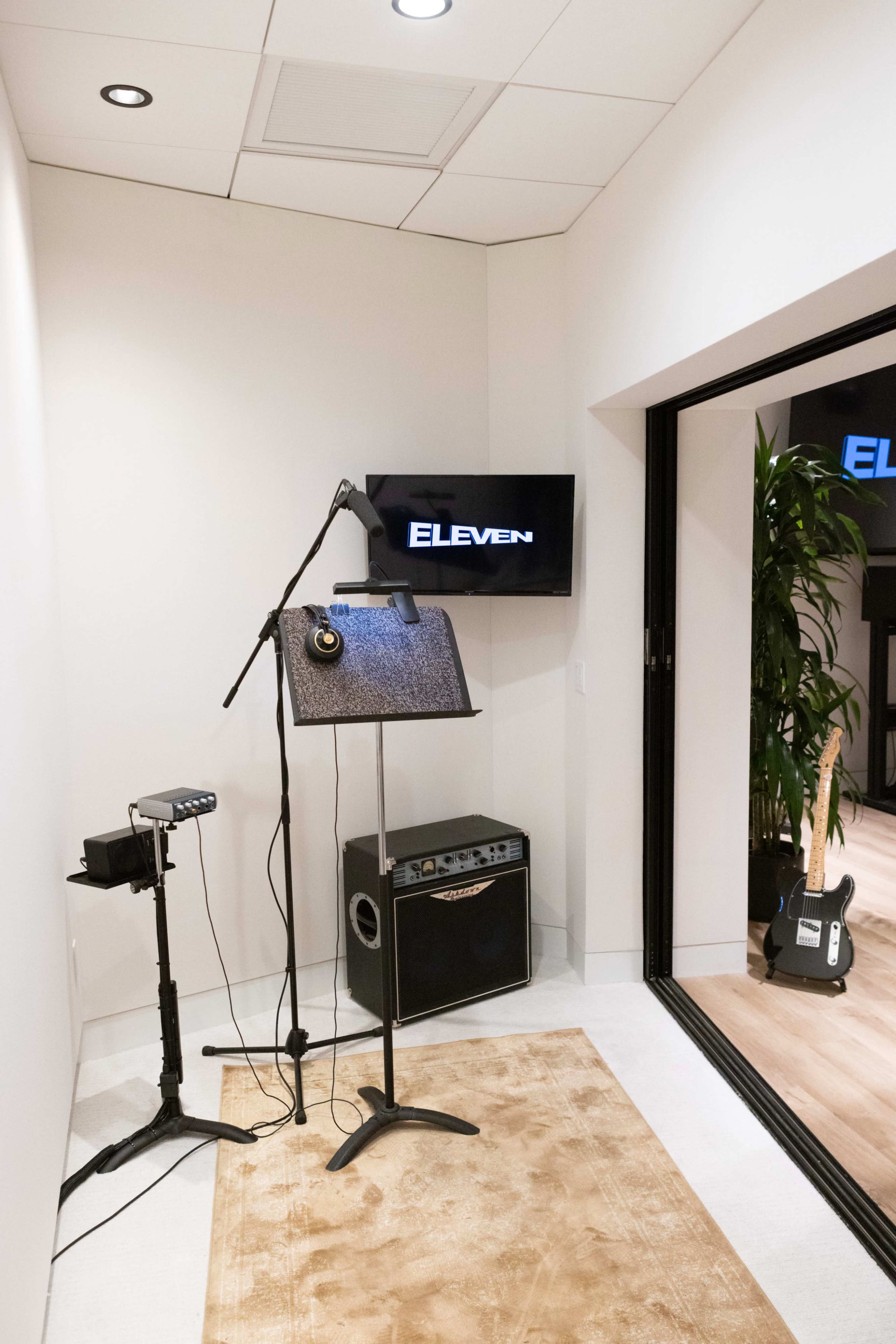 A small recording studio featuring a microphone on a stand, a music stand, an amplifier, and a television displaying the word "ELEVEN."