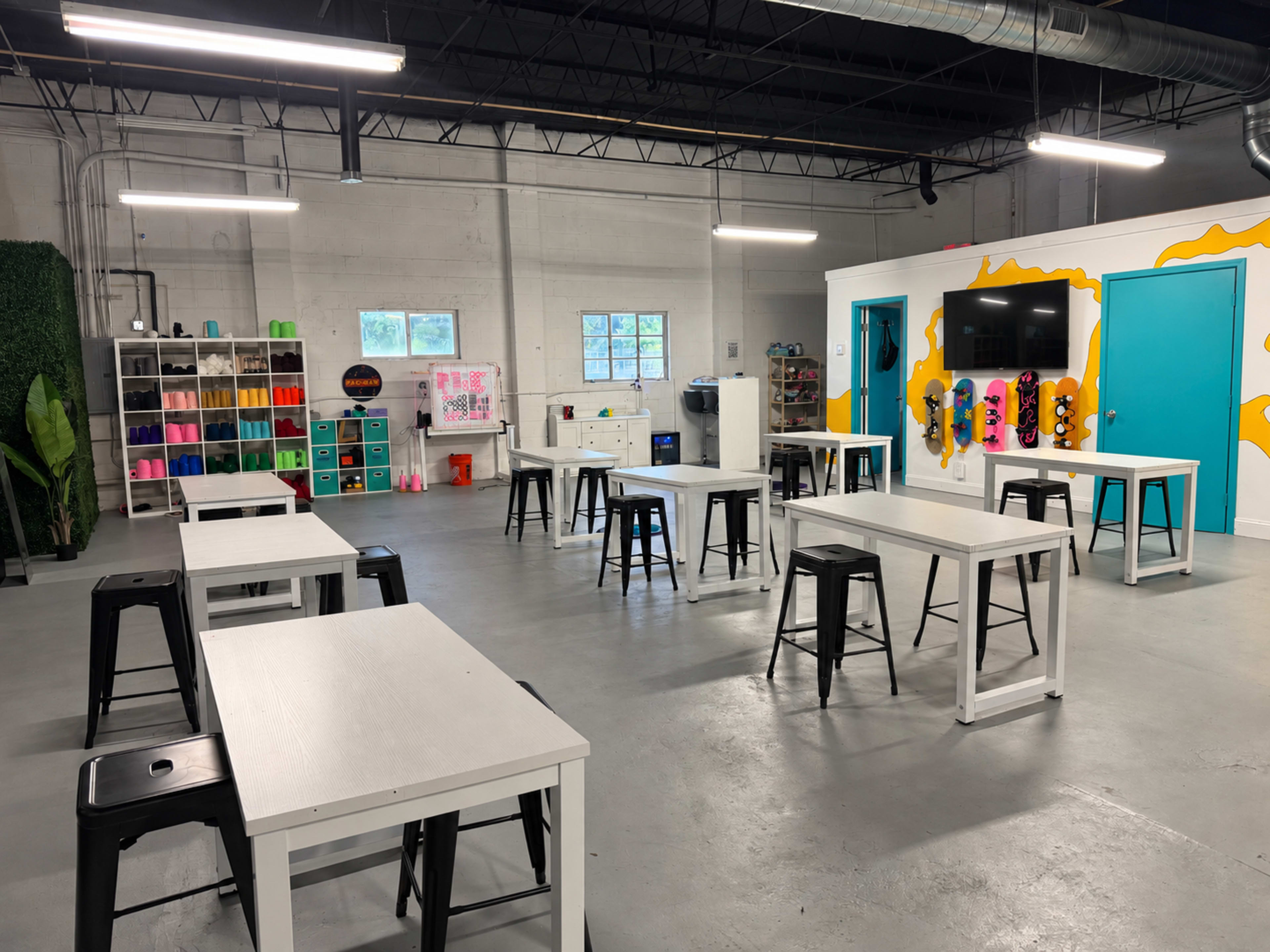 The image shows a spacious room with several white tables and black stools, colorful storage bins in the background, and a bright blue wall with a television and decorative elements.