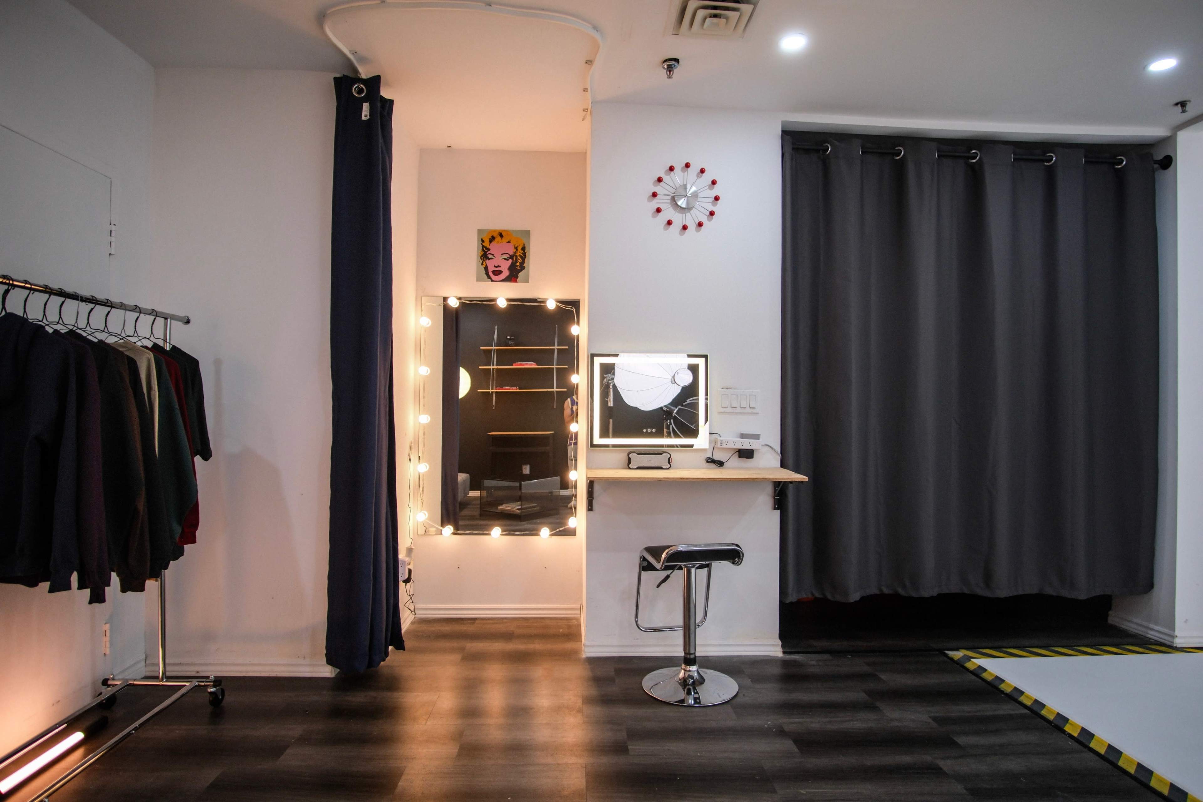 A well-lit dressing area featuring a mirror with lights, a small counter with a chair, and clothing racks along one wall.