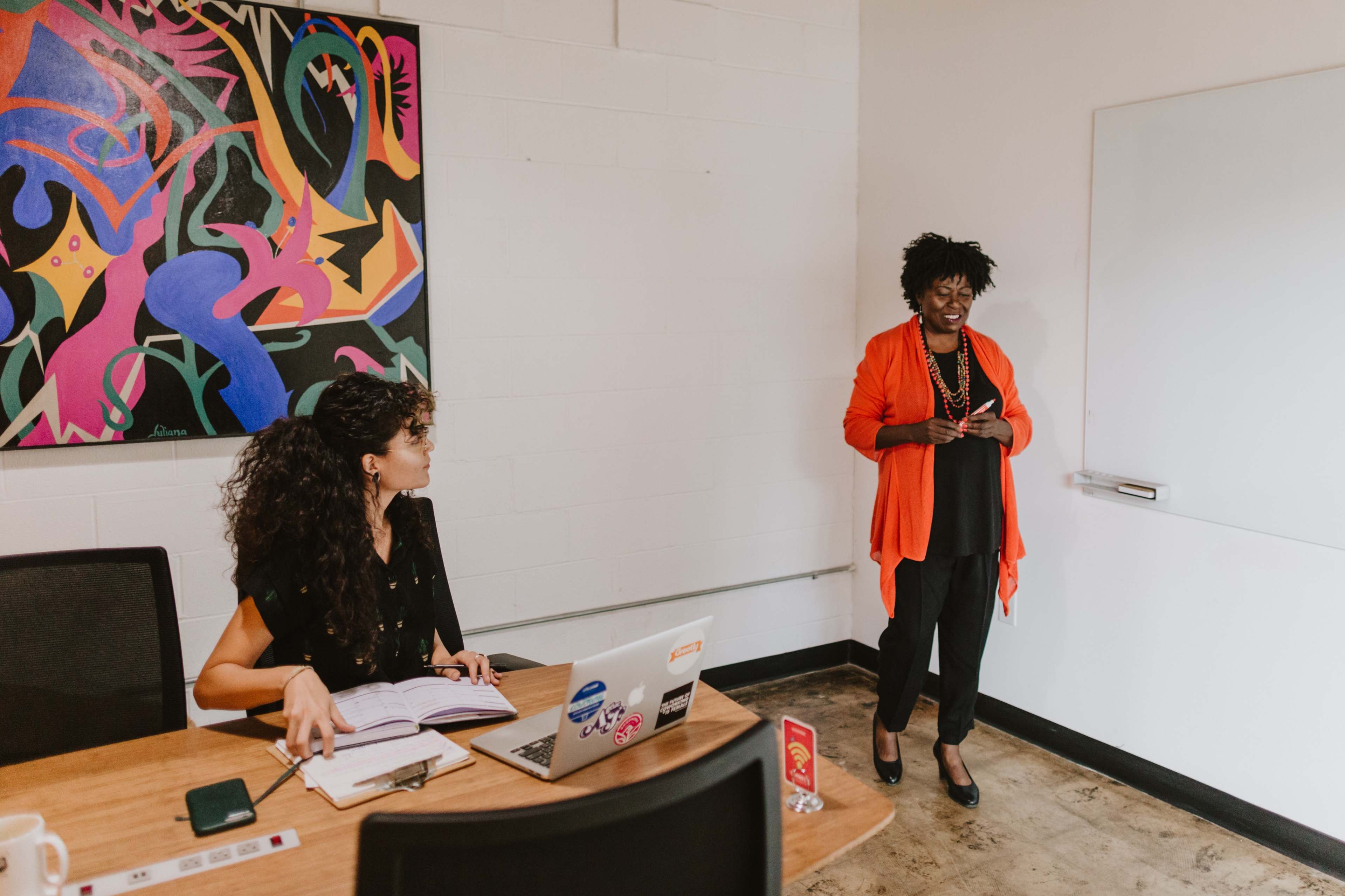 A woman in an orange blazer speaks to a seated woman at a table with a laptop, in a modern meeting room adorned with colorful artwork.