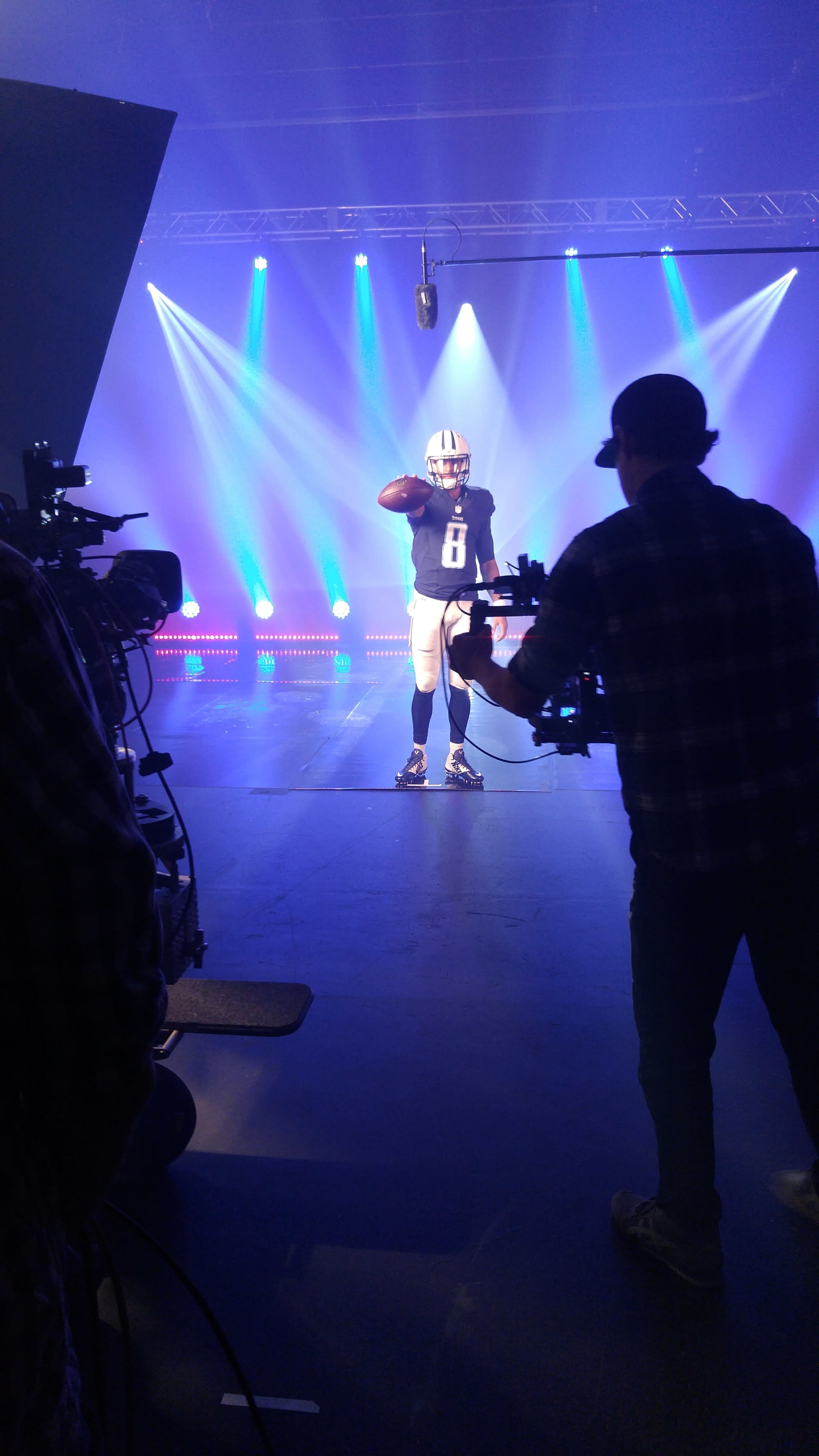 A football player poses in a studio under bright lights while a crew records the scene.