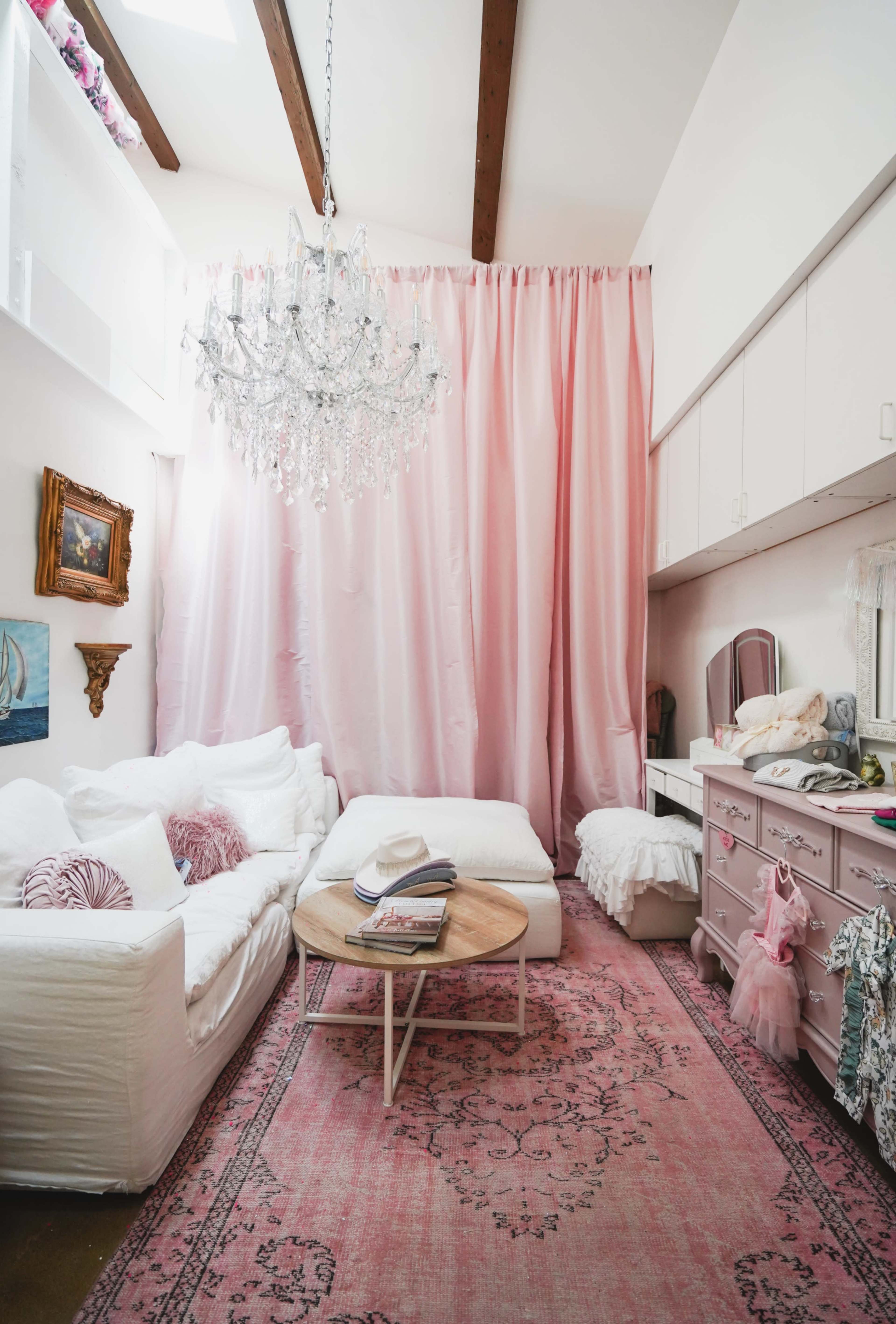 A cozy living room features a white sectional sofa, a circular coffee table, a pink area rug, and a chandelier, with pink curtains and light-colored furniture.