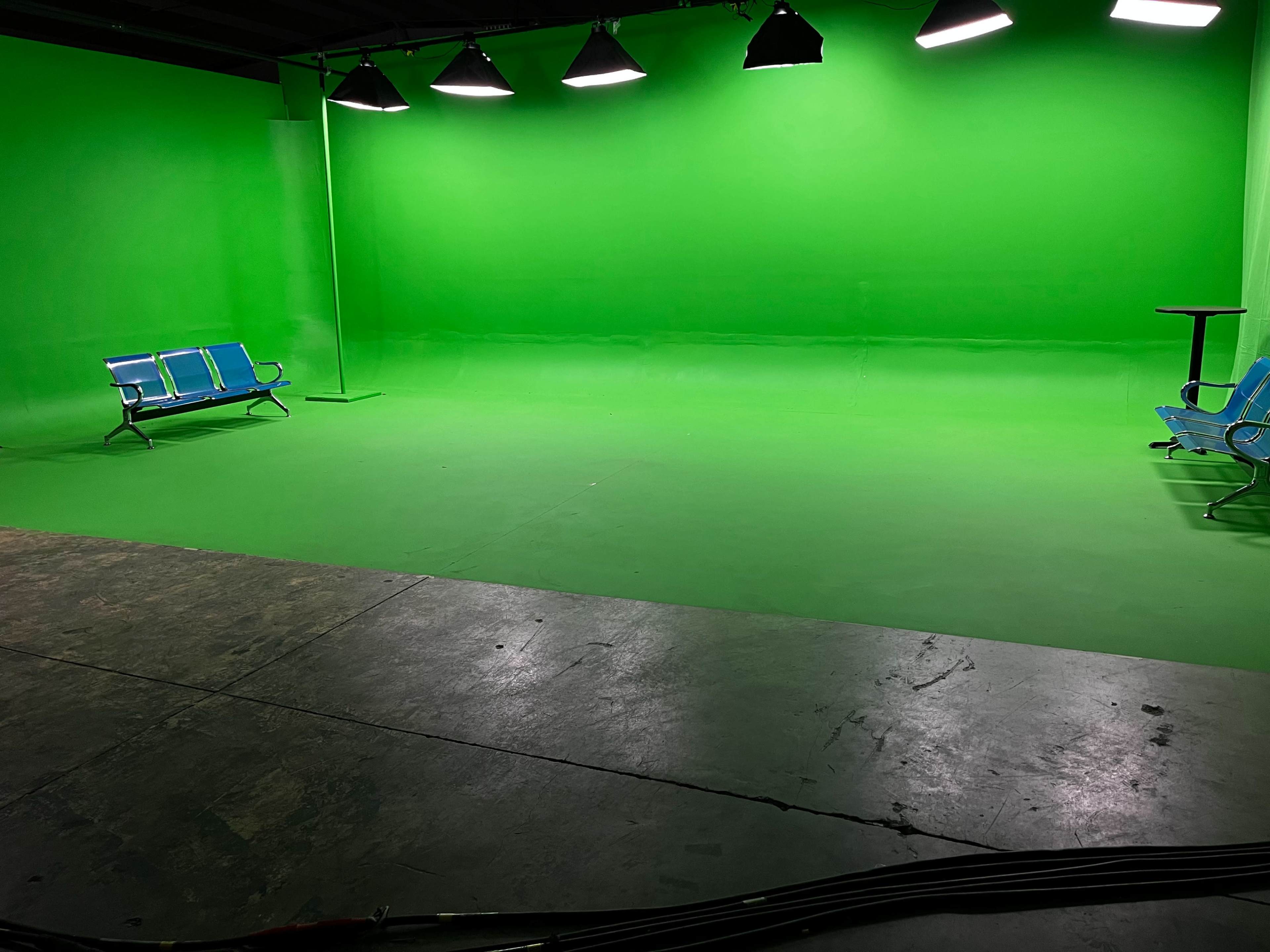 The image shows a green screen studio featuring two blue benches and a small table on a concrete floor.