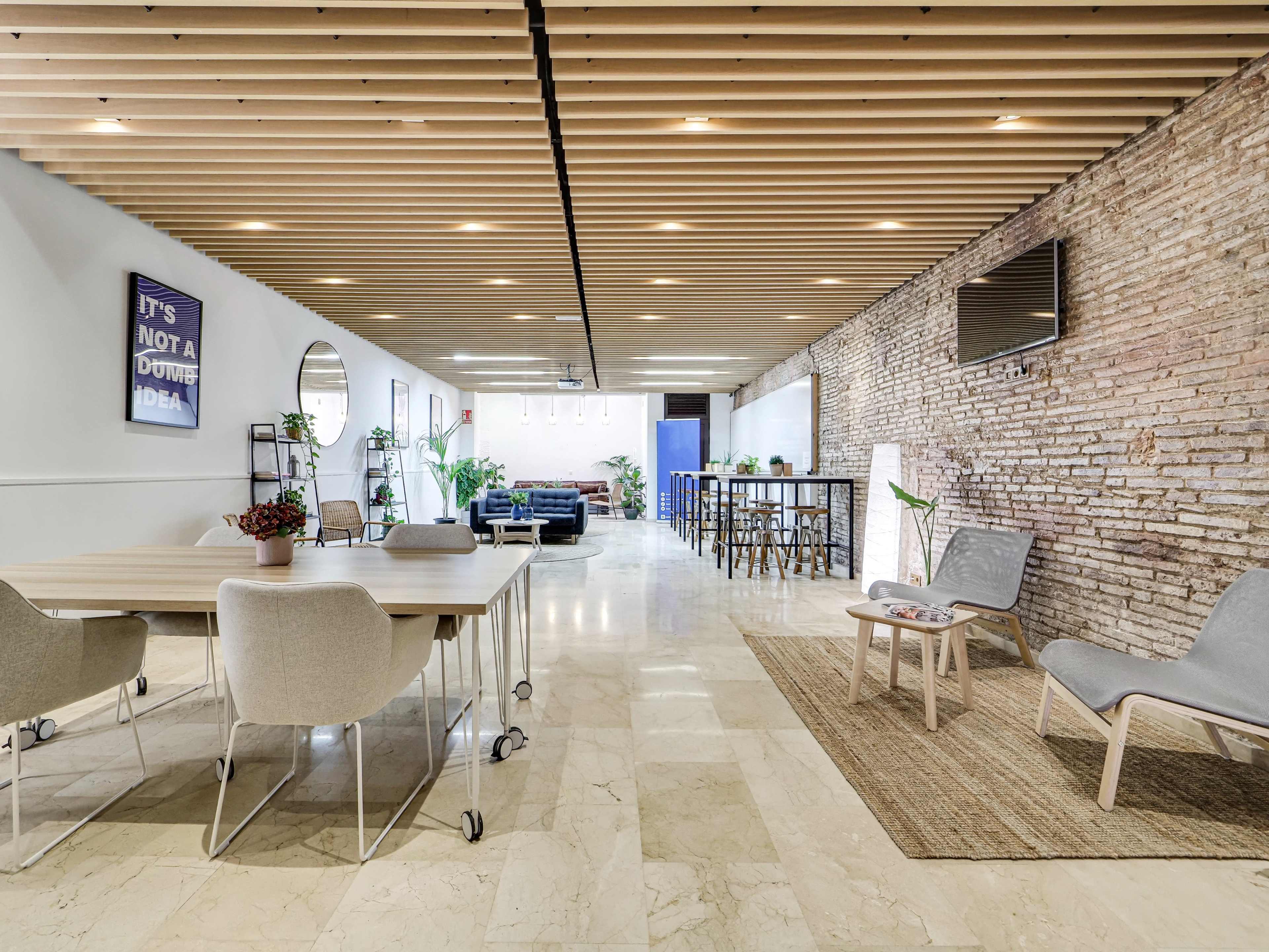 The image shows a modern open-concept workspace featuring wooden beams, a mix of seating arrangements, and a large brick wall.