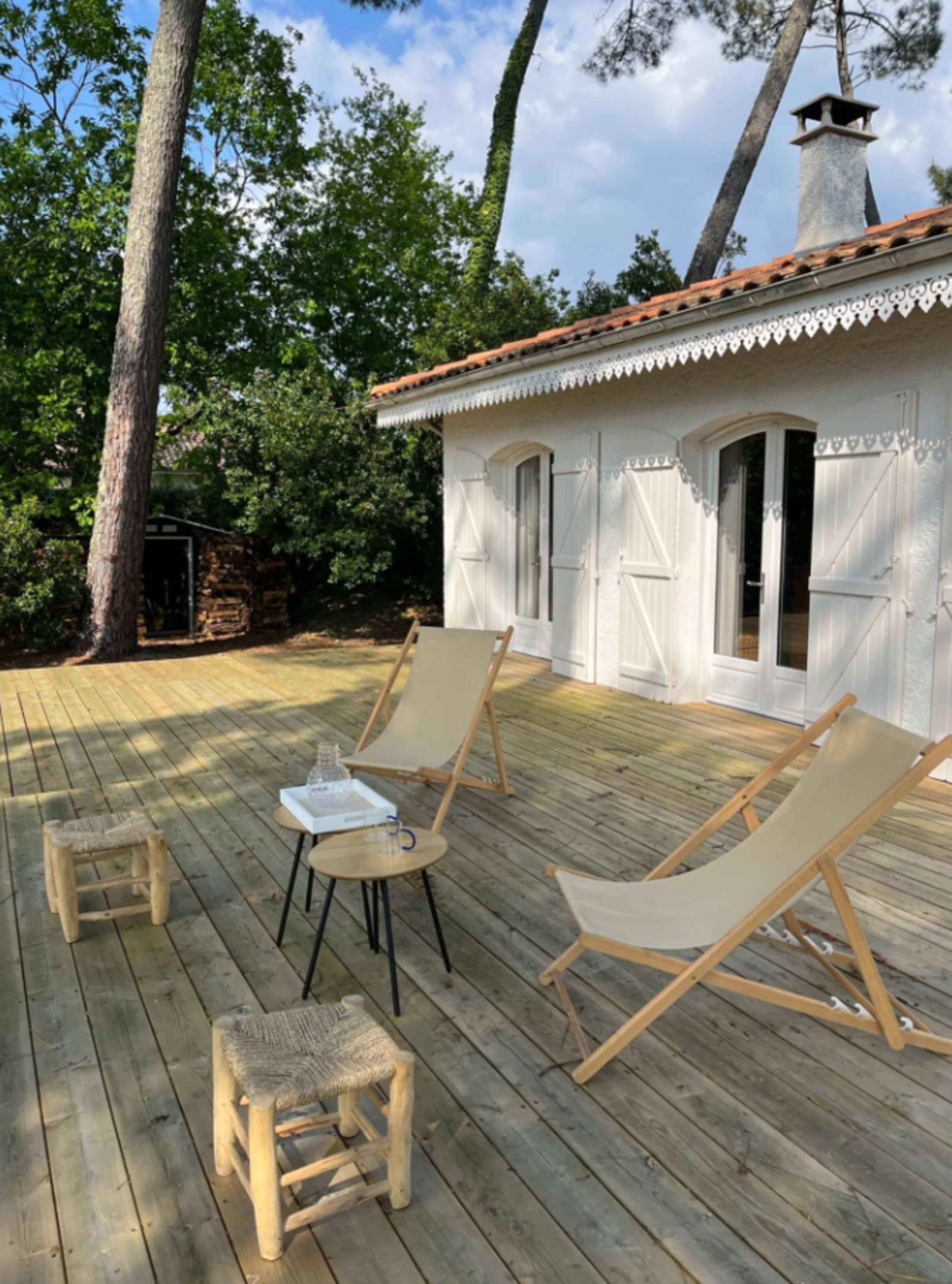 The image shows a wooden deck with two lounge chairs, a small table, and a house with white shutters surrounded by trees.
