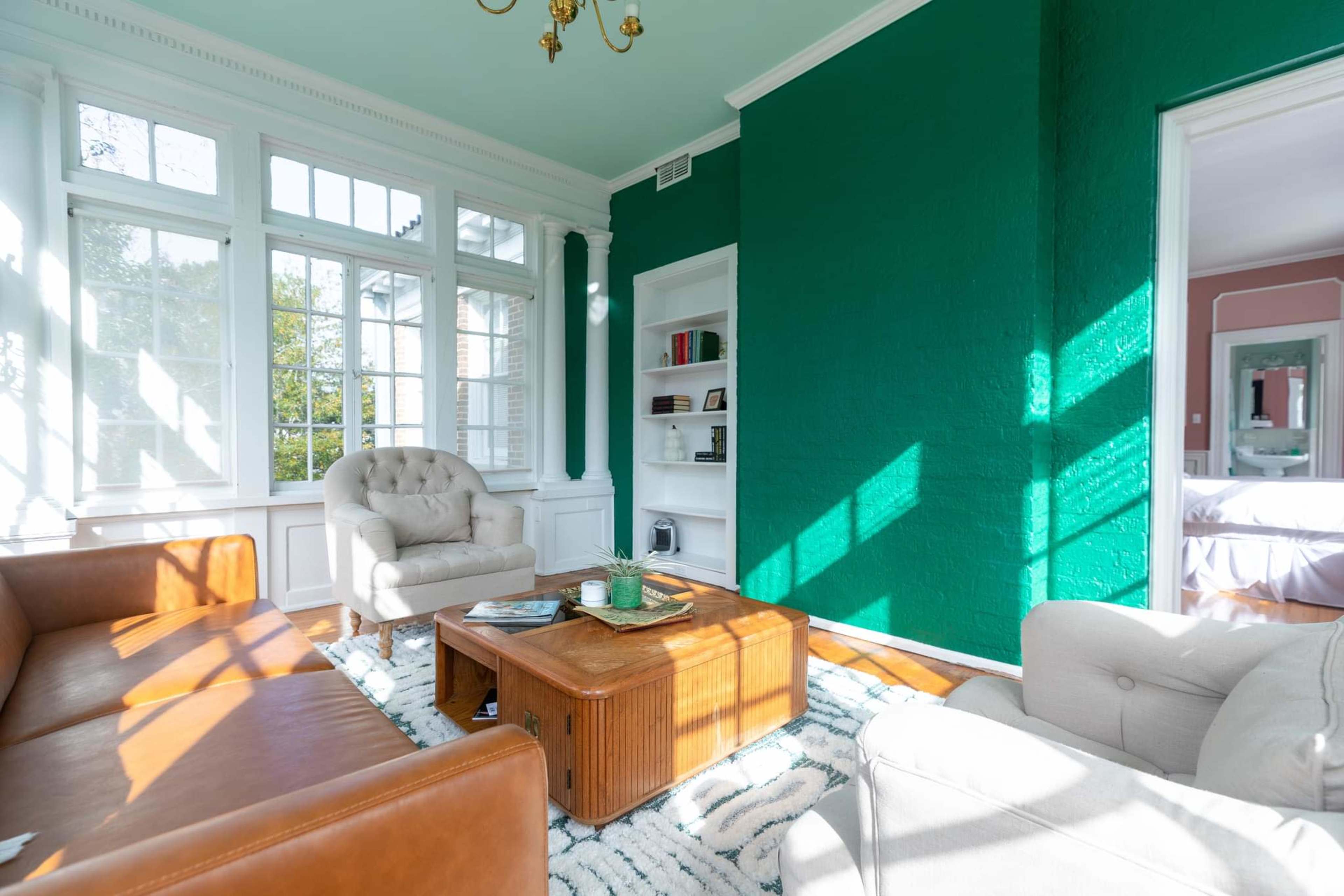 The room features a green accent wall, large windows allowing sunlight to stream in, and a mix of leather and fabric seating around a wooden coffee table.