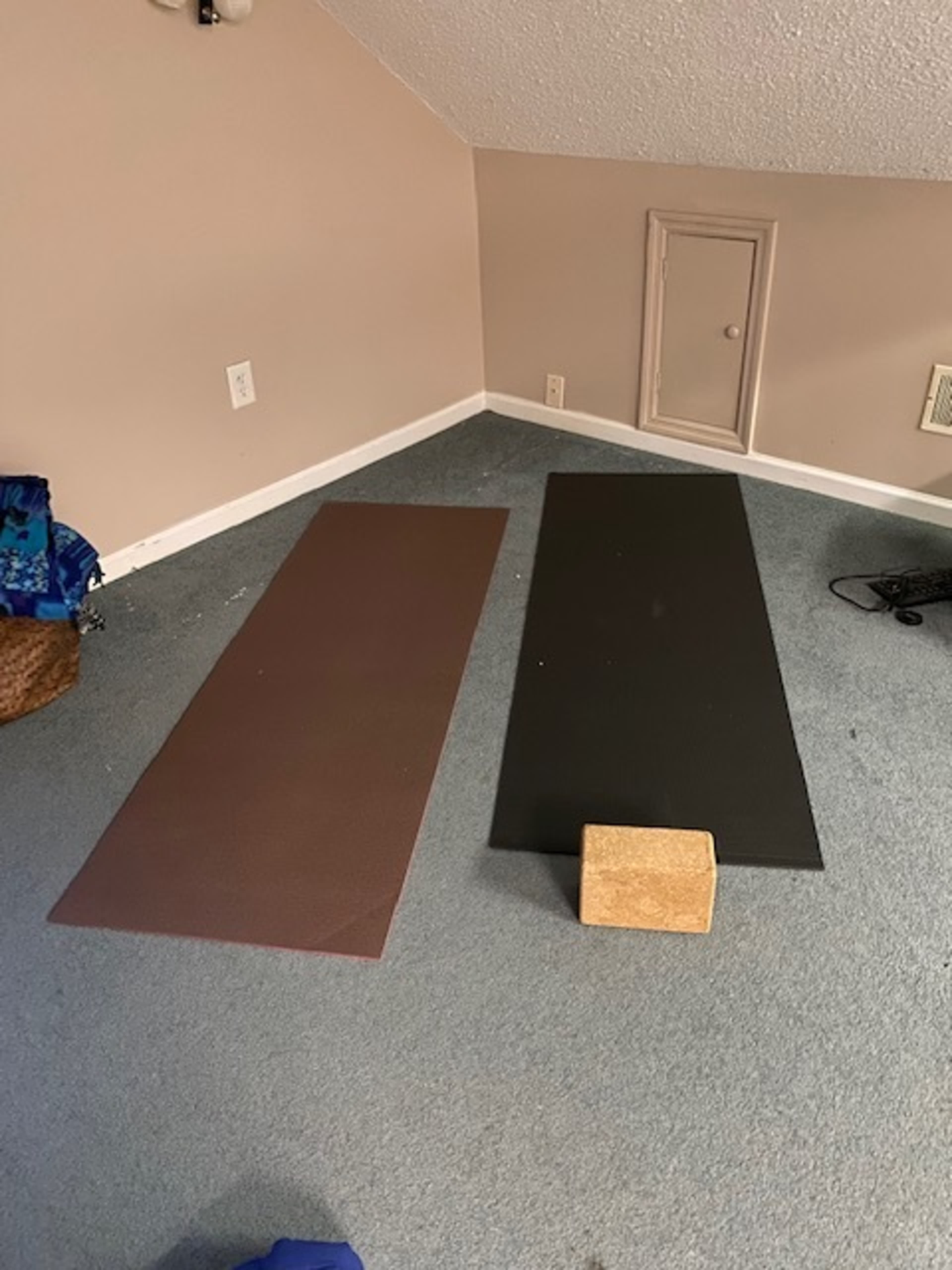 Two yoga mats, one brown and one black, are placed side by side on a light blue carpeted floor, with a wooden yoga block positioned nearby.