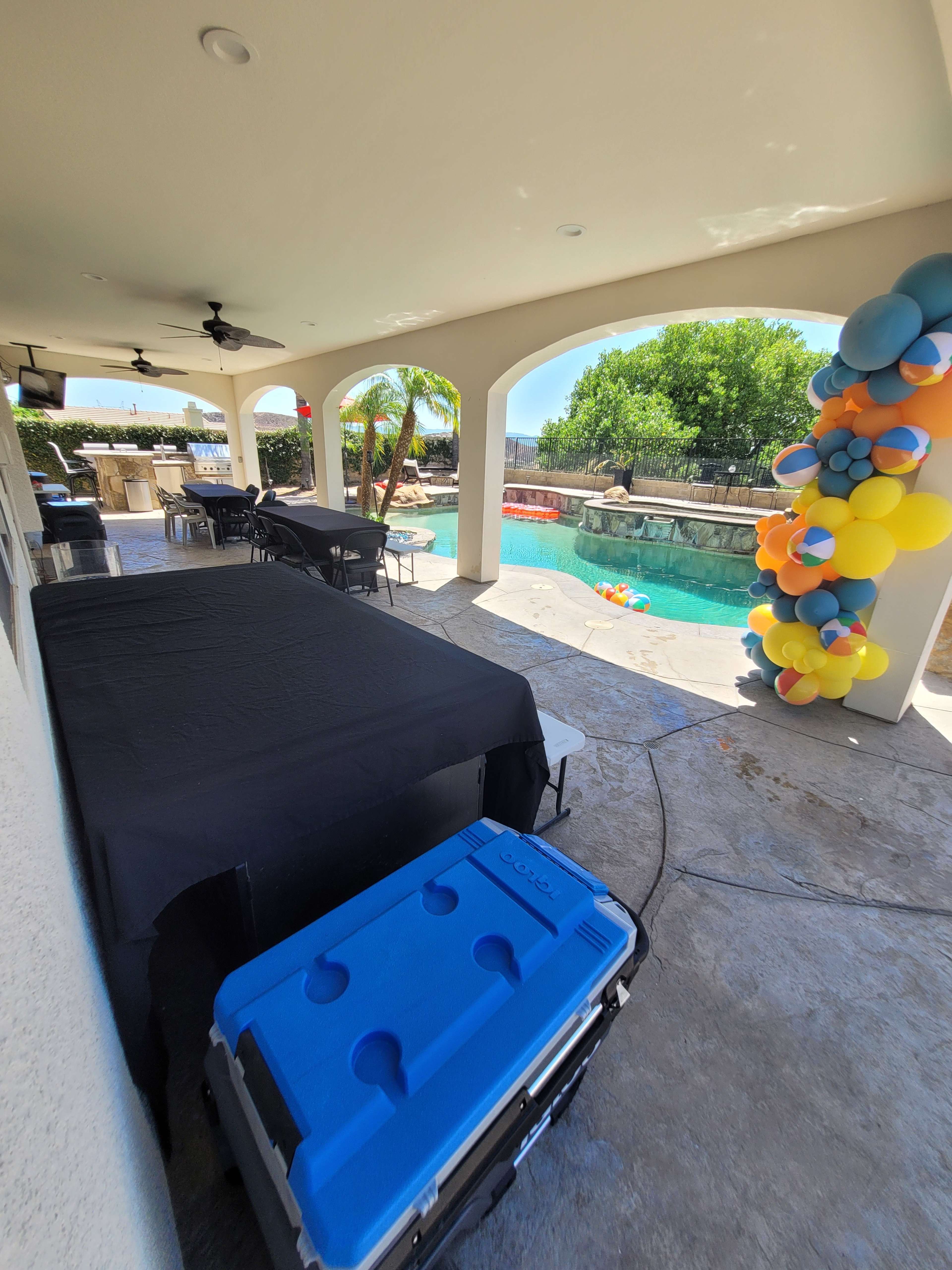A covered outdoor patio area features a table set up with a blue cooler and colorful balloons near a swimming pool.