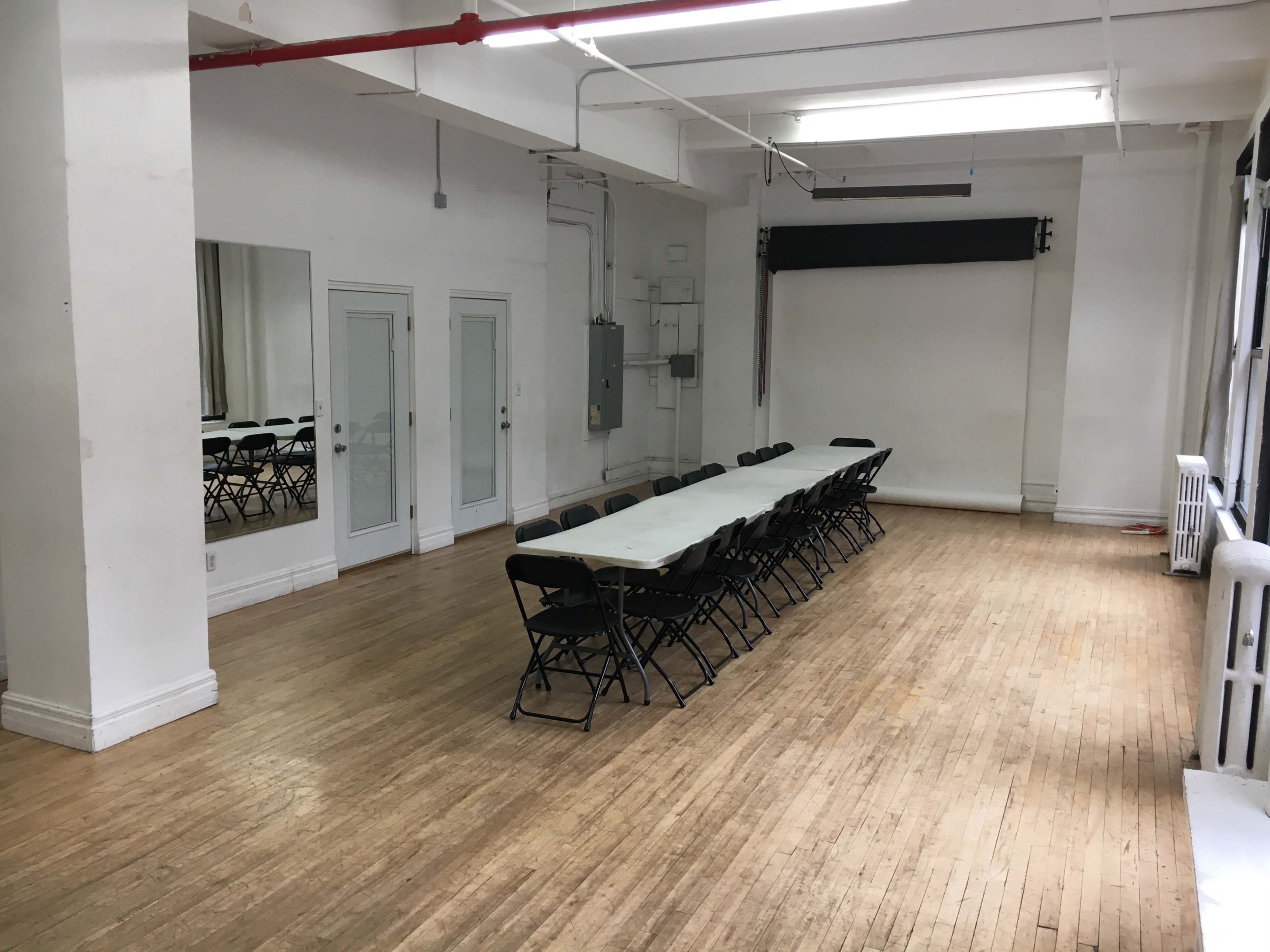 A long rectangular table with chairs is arranged in an empty room featuring white walls and wooden flooring.