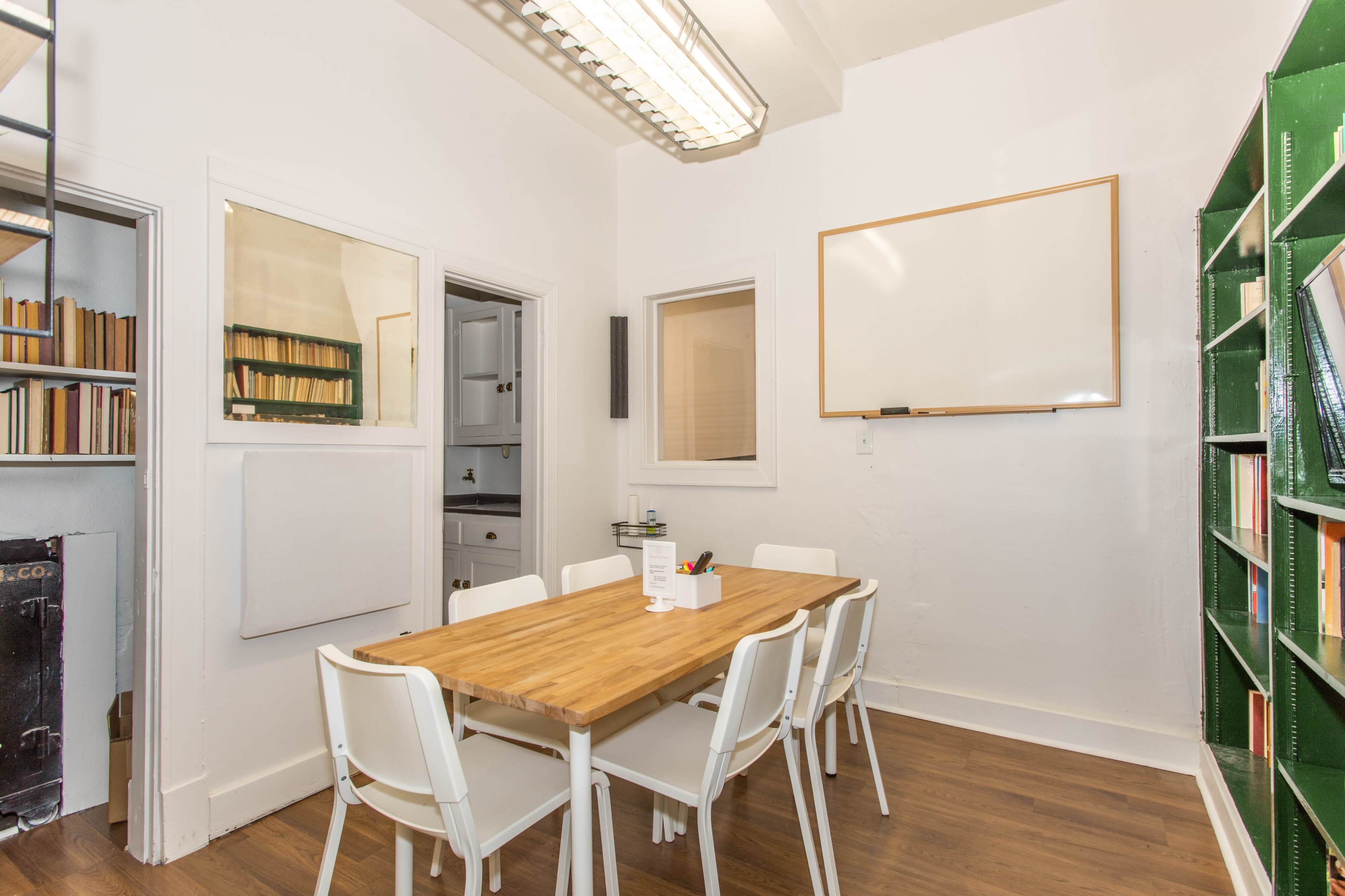 A small meeting room features a wooden table surrounded by six white chairs, with bookshelves and a whiteboard on the walls.