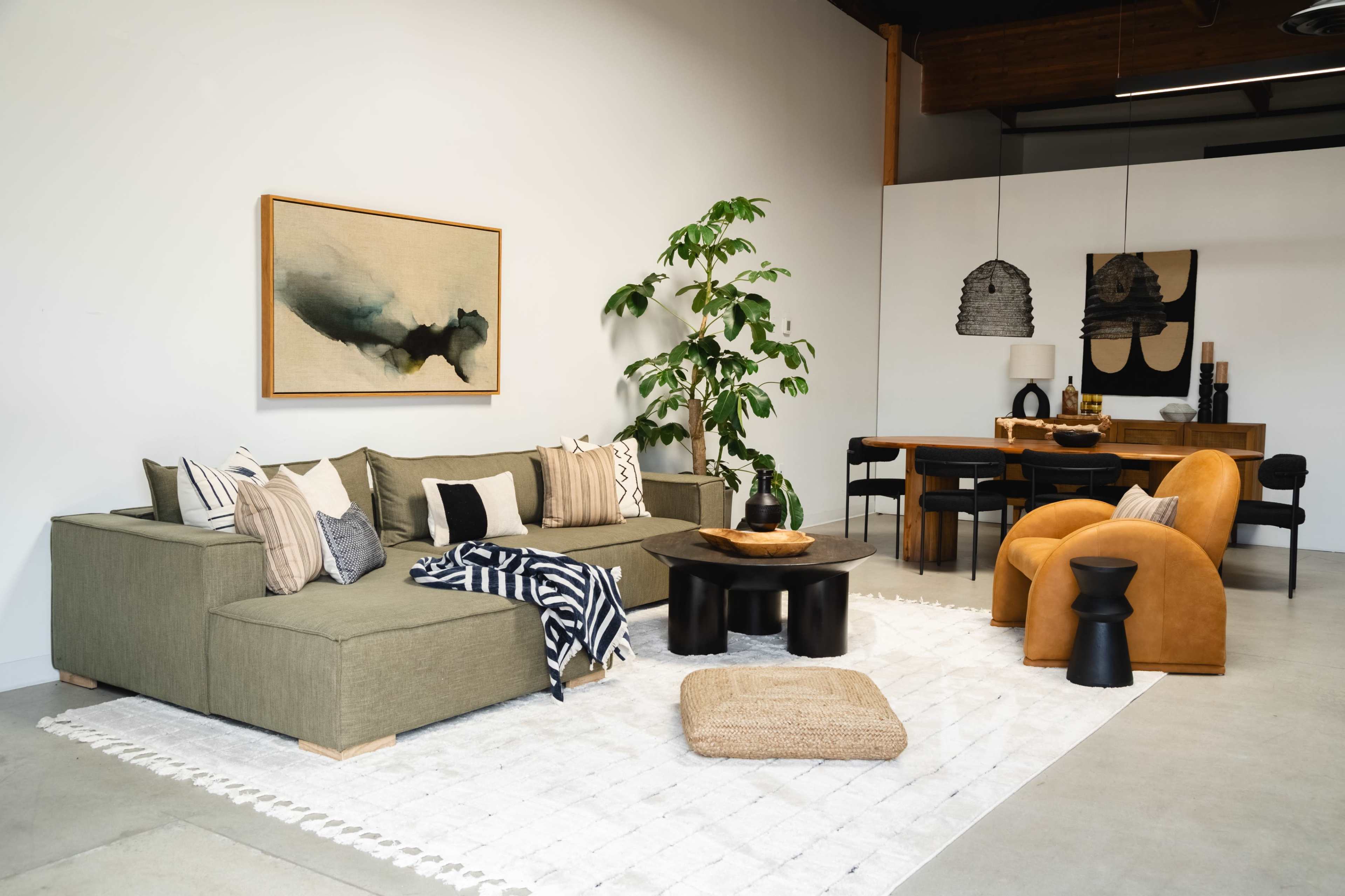 A modern living room featuring a sectional sofa, a large artwork on the wall, a dining area with a wooden table and black chairs, and decorative plants.