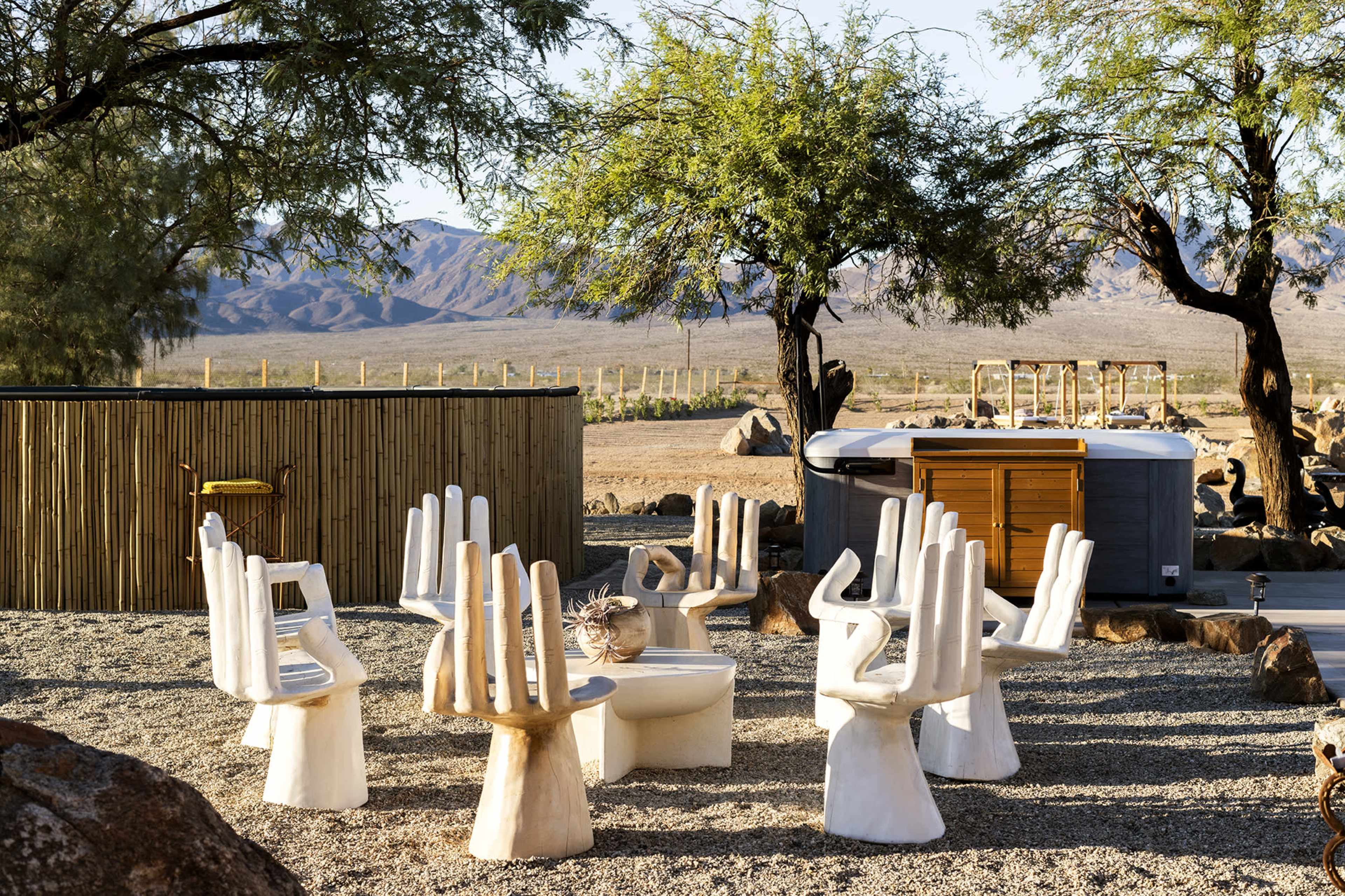 The scene features a unique outdoor seating area with sculptural chairs resembling hands arranged around a circular table, surrounded by desert landscape and trees.